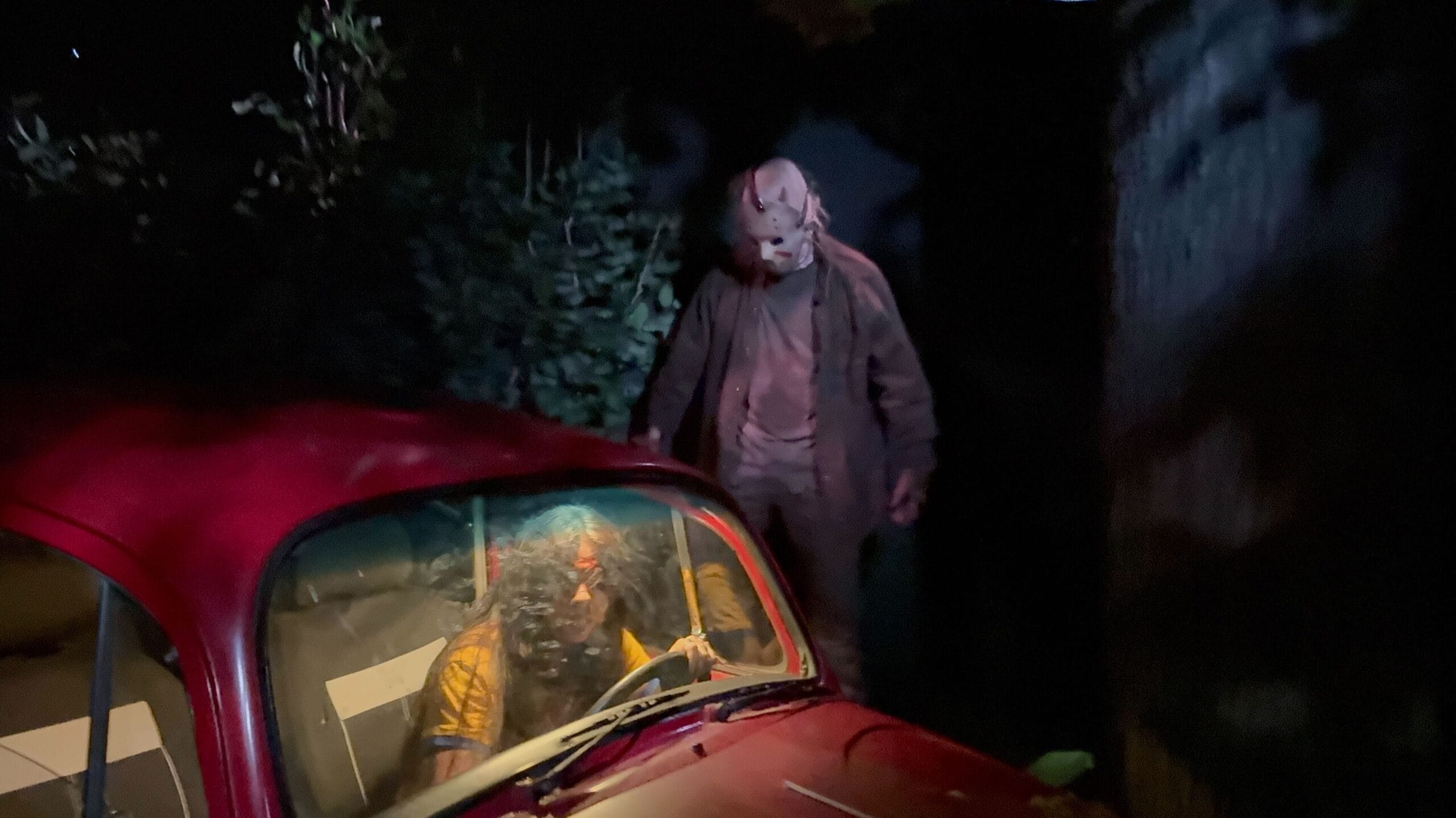 A masked figure stands behind a person in a yellow shirt sitting in a red car at night.