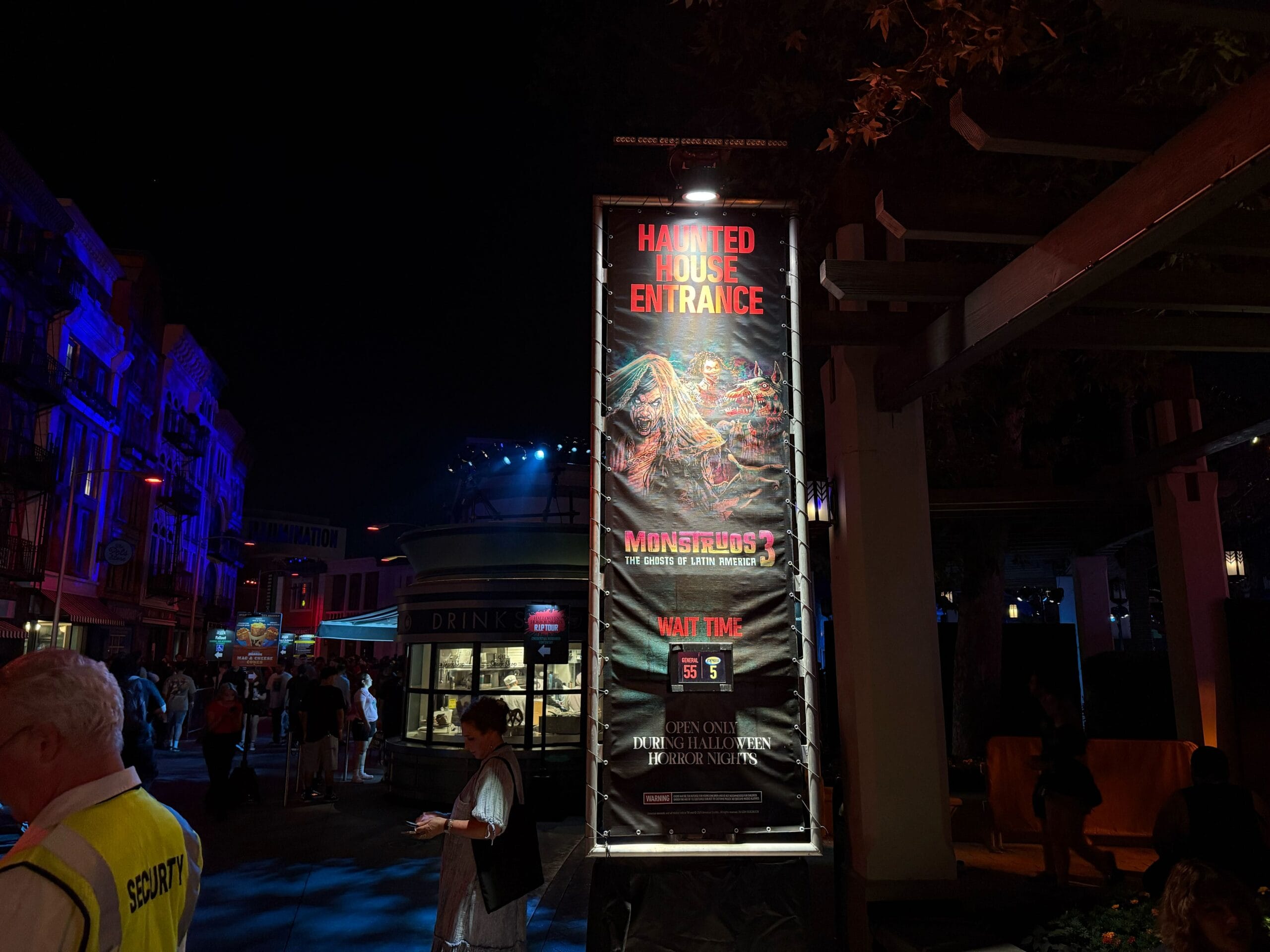 A lit sign at the HHN 2025 haunted house entrance shows a 55-minute wait; people and USH security stand nearby.