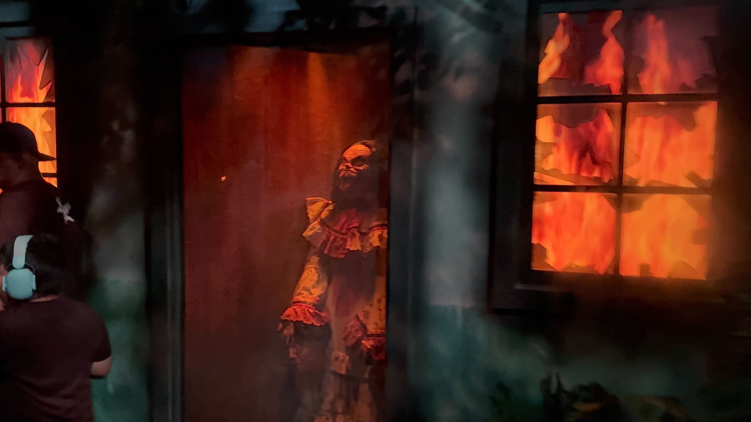 A sinister clown from Monstros 3 stands in a doorway near burning windows, people nearby in a dark, smoky scene.