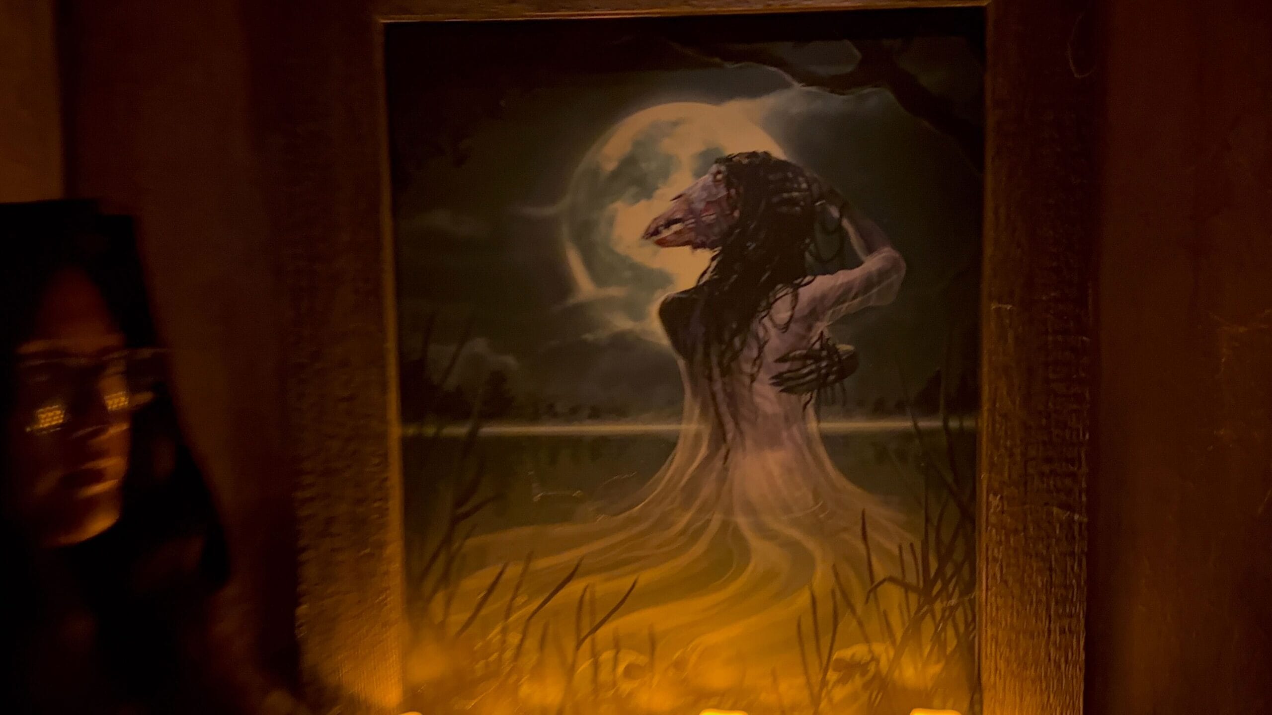A painting of a wolf-headed woman in white, embraced by shadow—Monstros 3 at USH under the full moon.