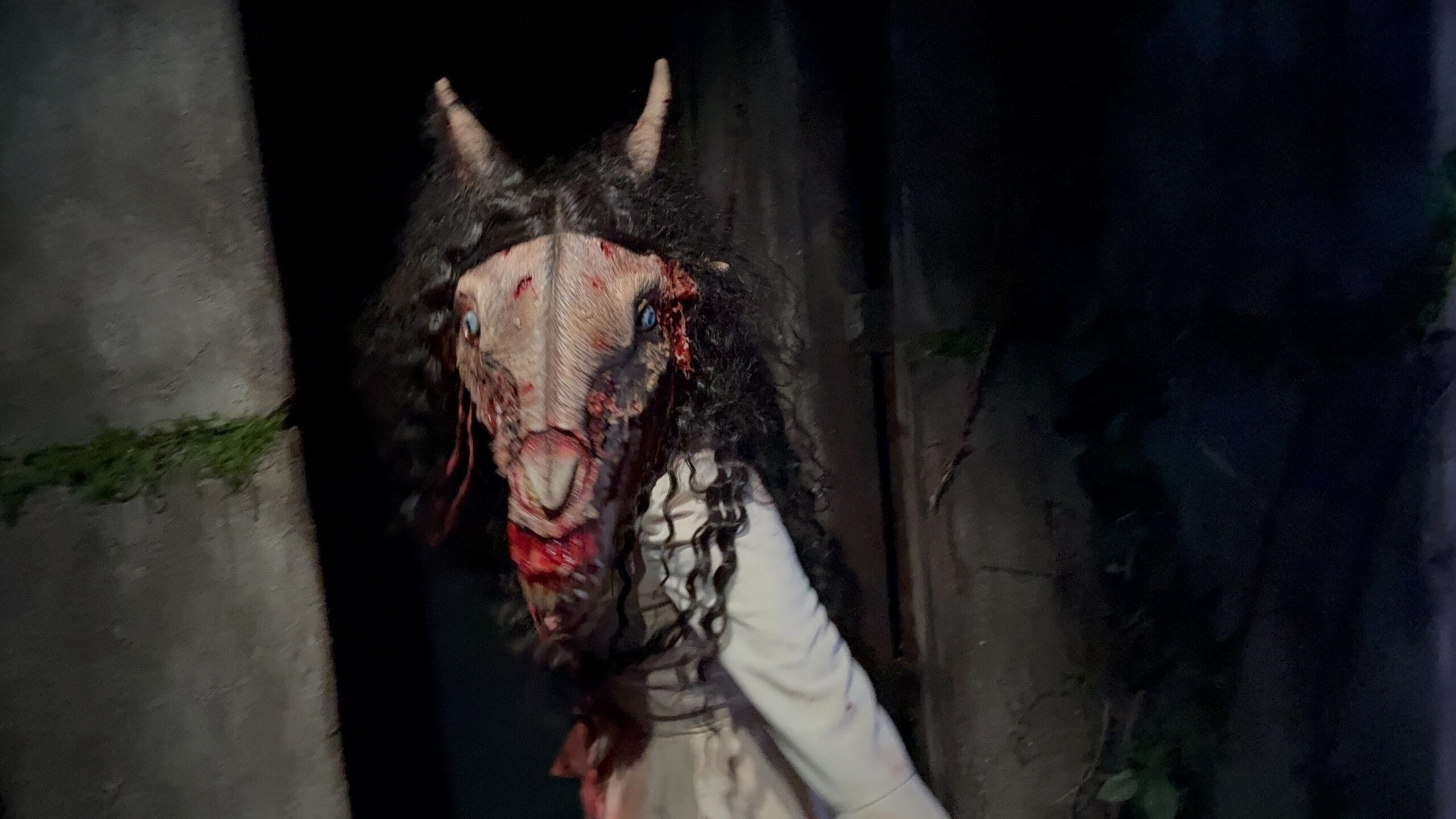 A person in a creepy, horned Monstros 3 mask with long black hair haunts a dark, eerie setting at USH.
