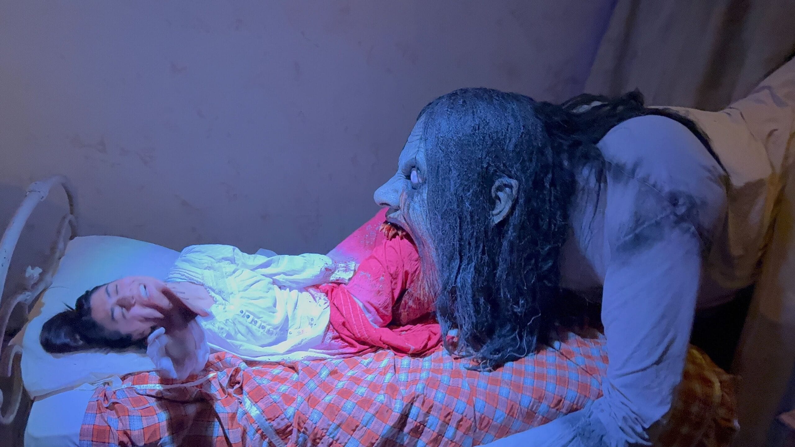 A scary creature from Monstros 3 leans over a terrified woman lying in bed with blood on her dress.