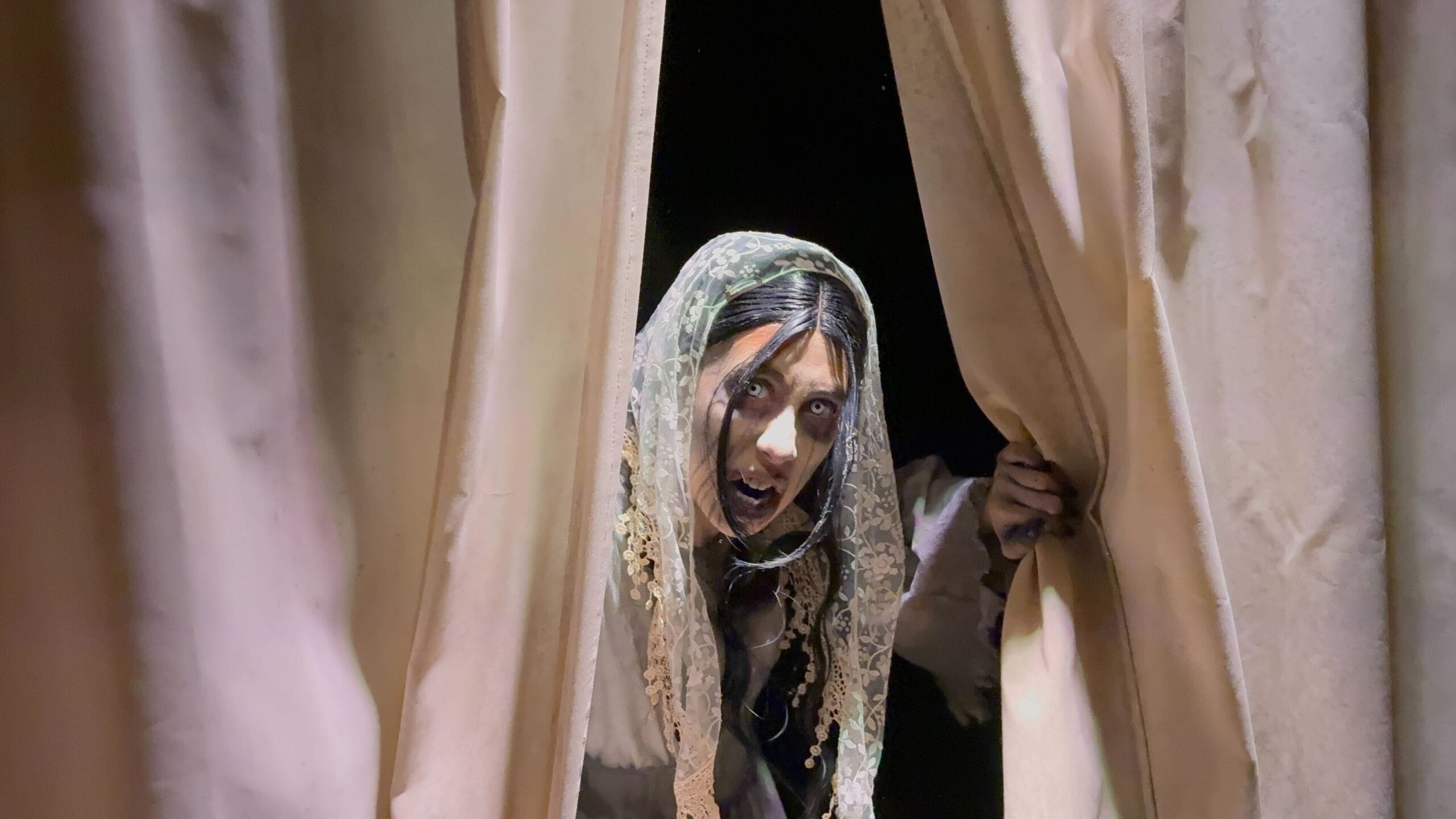 A person in a lace veil peers through parted beige curtains at USH, channeling dramatic HHN 2025 intensity.