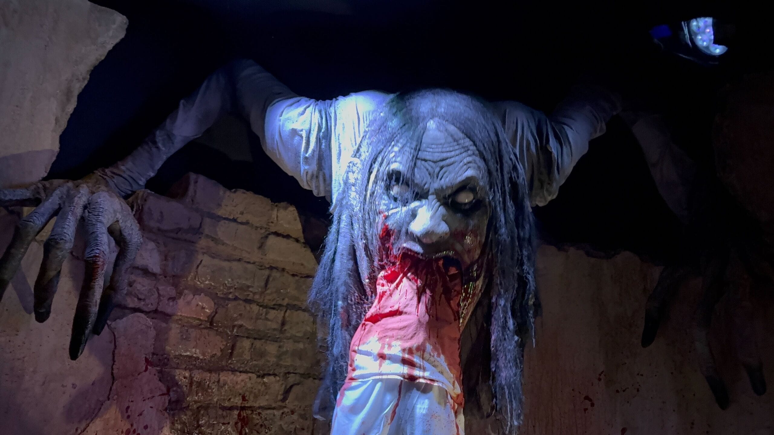 A creepy Monstros 3 creature with long hair bites a bloody arm in a dark, eerie setting at HHN 2025.