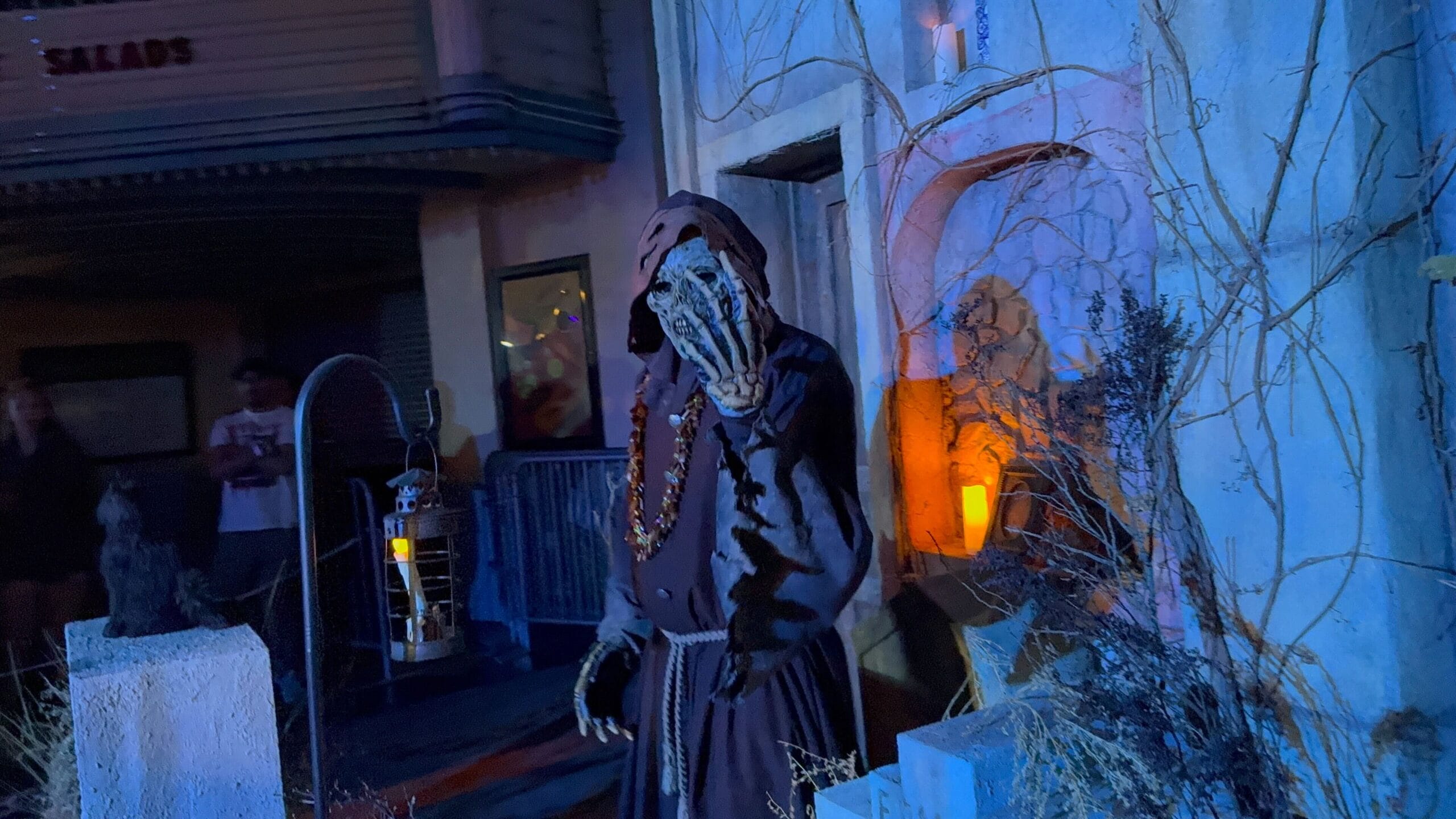 A creepy hooded figure in a skeleton mask looms by a lantern at USH during Monstros 3 at HHN 2025.