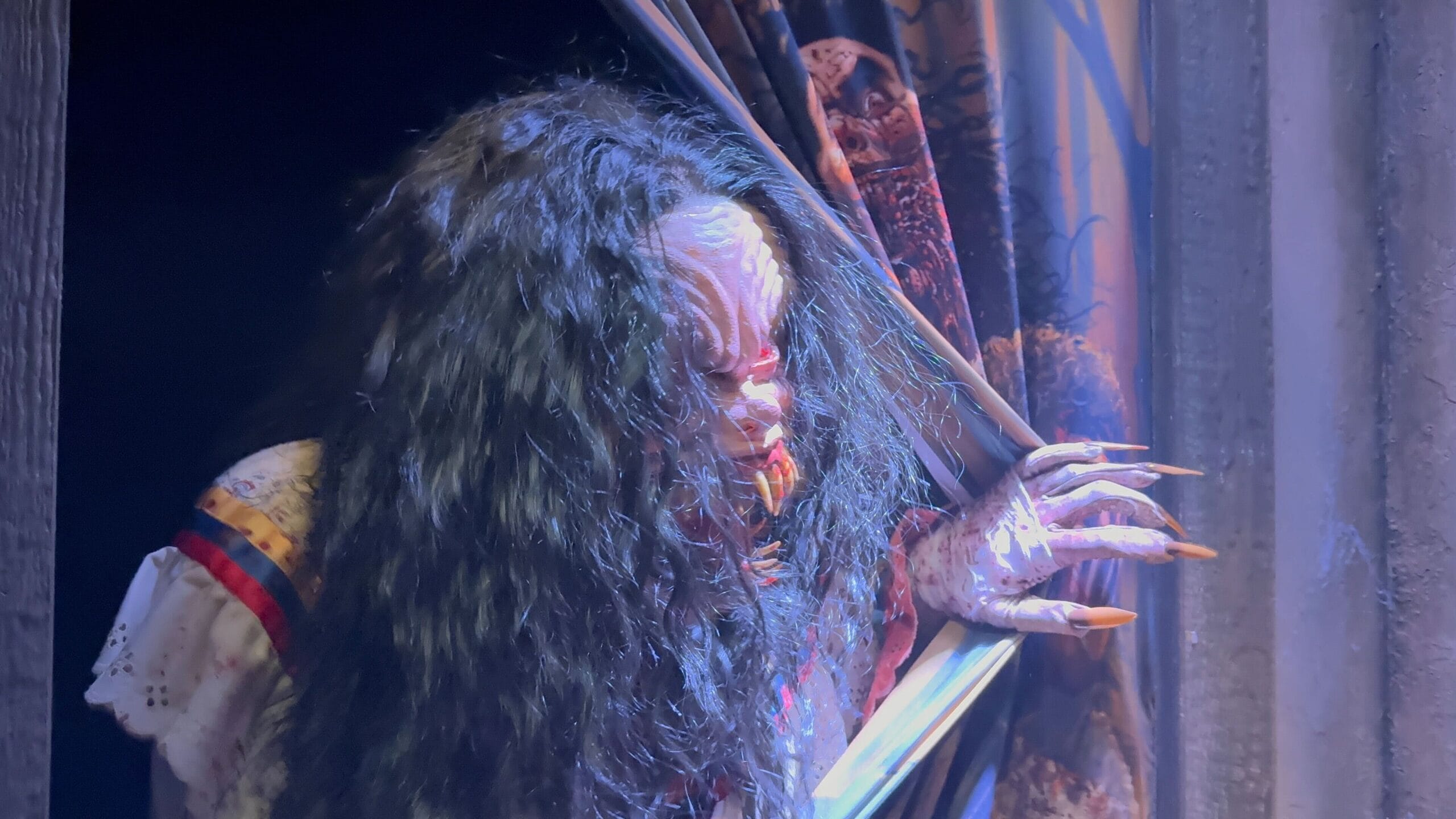 A creepy figure with long dark hair and claws peers through torn curtains at HHN 2025's Monstros 3, bathed in eerie lighting.