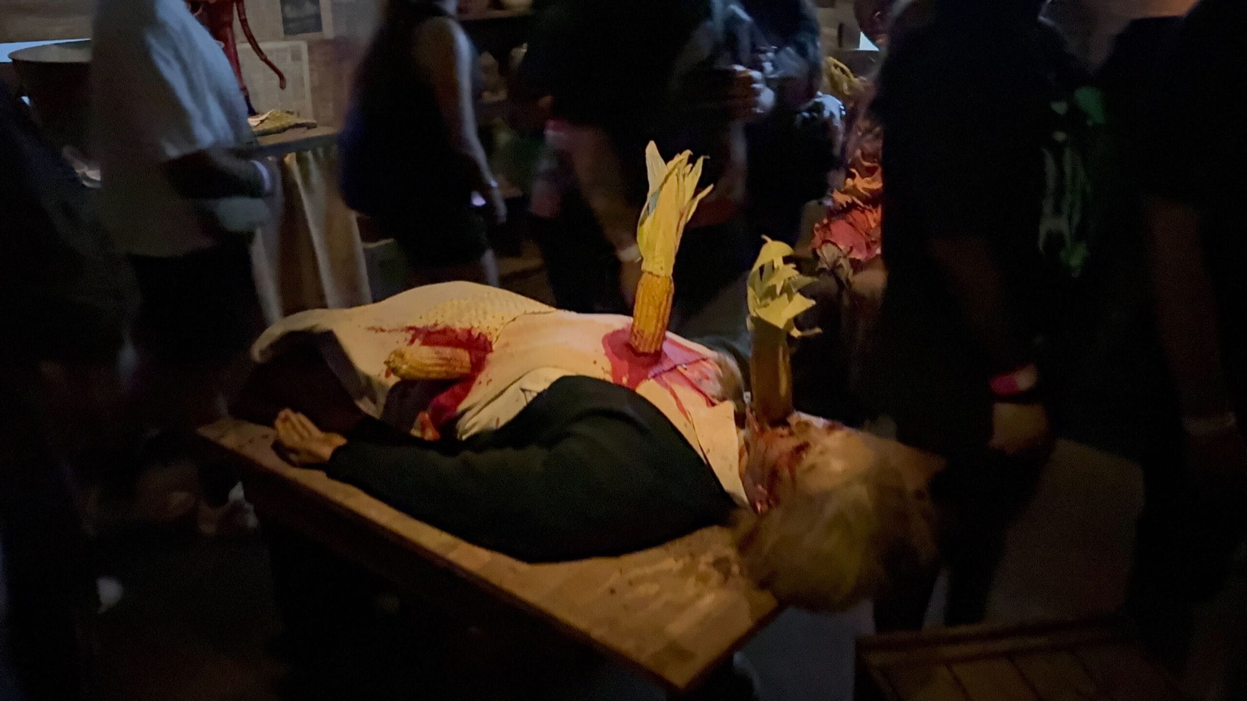 Un maniquí en una mesa con mazas de maíz falsas y sangre, rodeado de personas en un entorno de casa oscuro y embrujado.