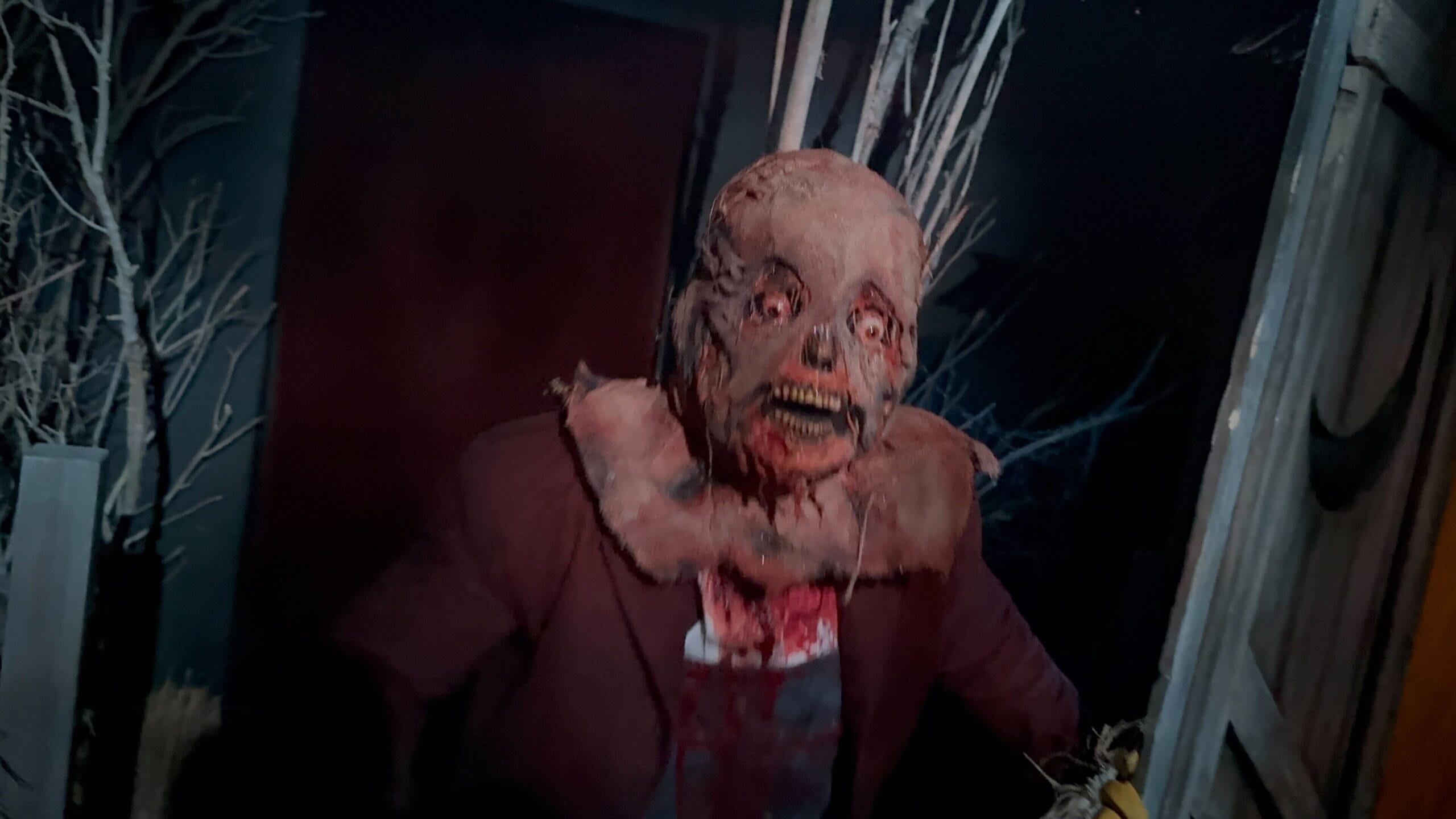 Una persona con un grotesco y sangriento disfraz zombie se encuentra en un entorno oscuro y misterioso con árboles desnudos.
