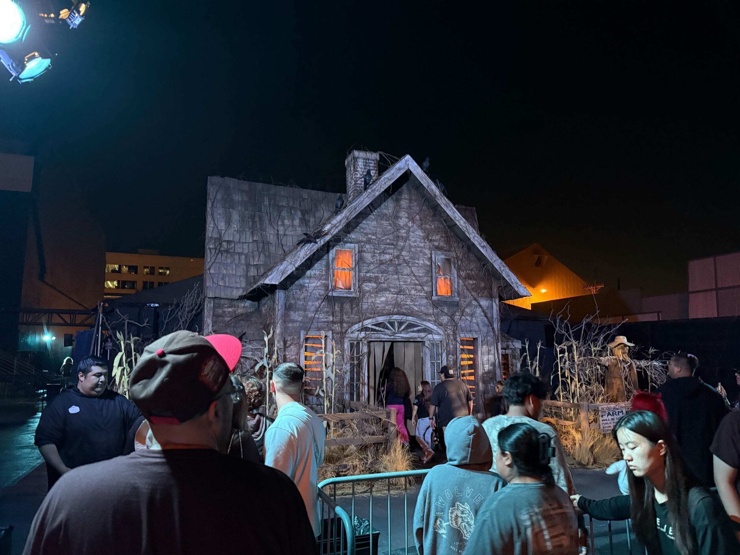 La gente espera en la fila por la noche afuera de una atracción de casa embrujada con iluminación misteriosa y árboles sin hojas.