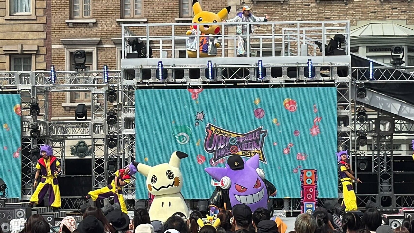 Pokémon mascots and dancers perform on stage at a Halloween Party, with Pikachu on a raised platform.