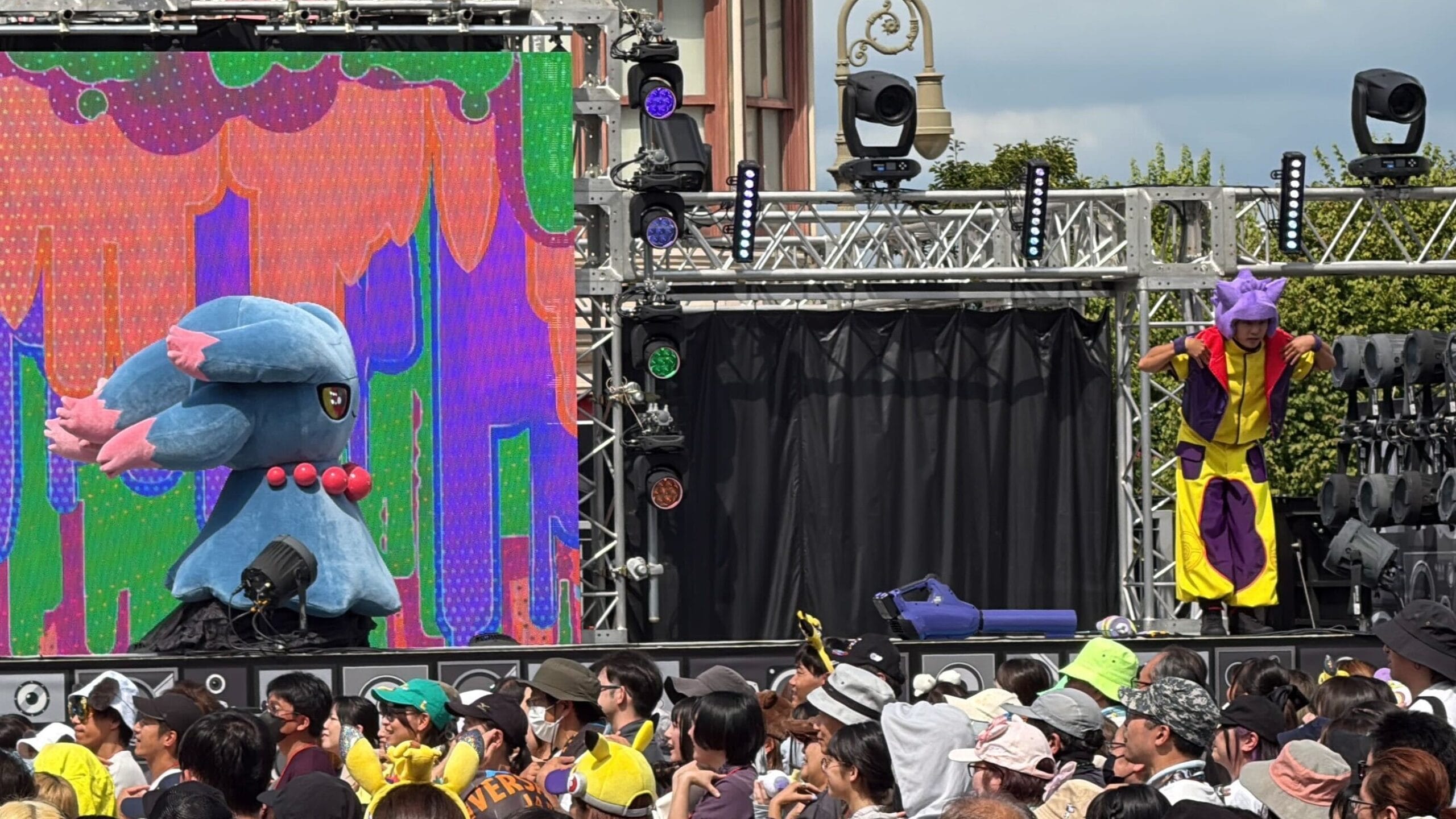 A person in costume and a large blue Pokémon perform on stage outdoors at a colorful Universal Studios Japan event.
