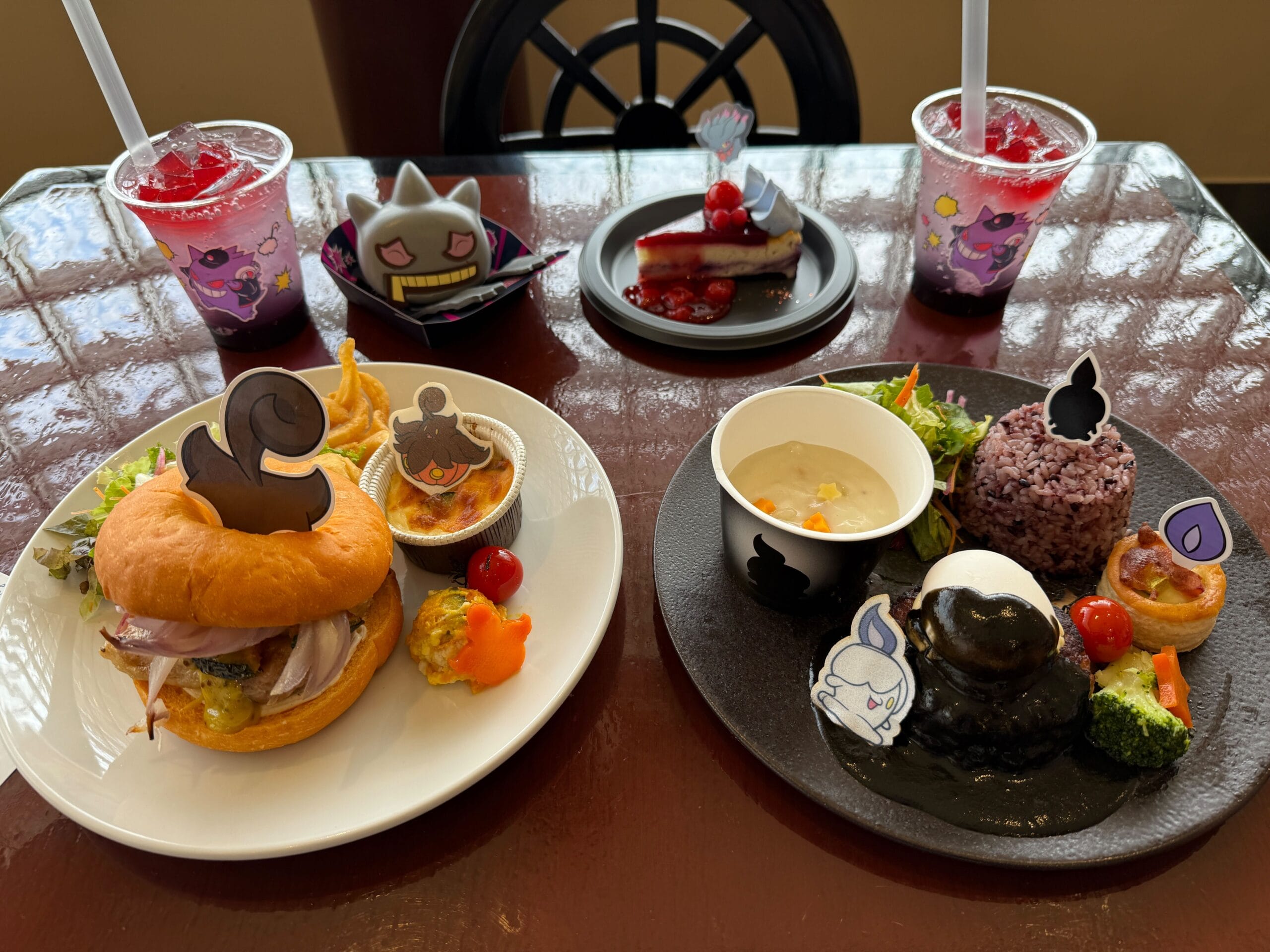 Two themed meals and drinks with ghost Pokémon decorations, plus a dessert plate, on a dark table near a window.