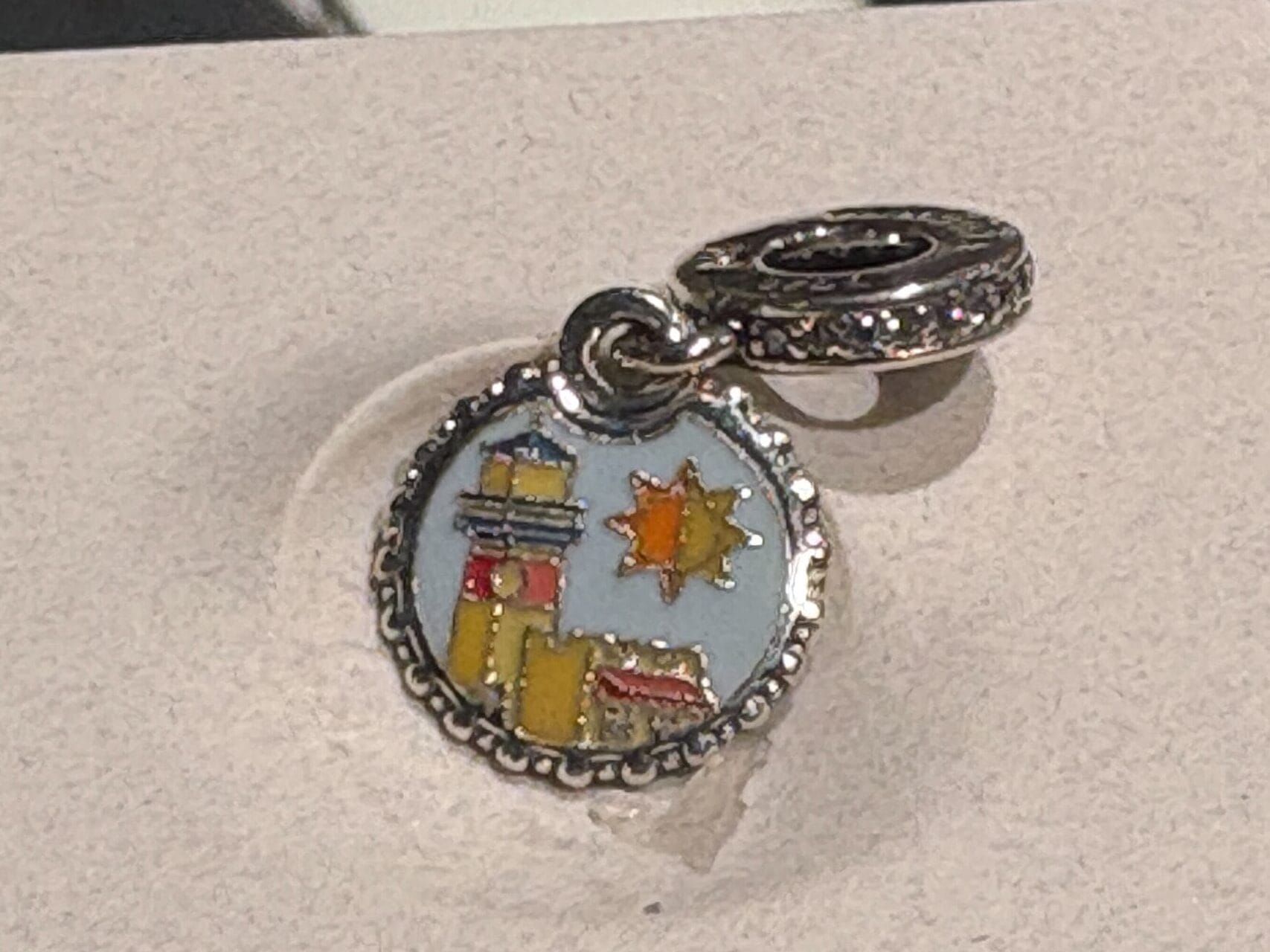 A silver charm with a colorful enamel cityscape and yellow sun design, displayed on a light surface.