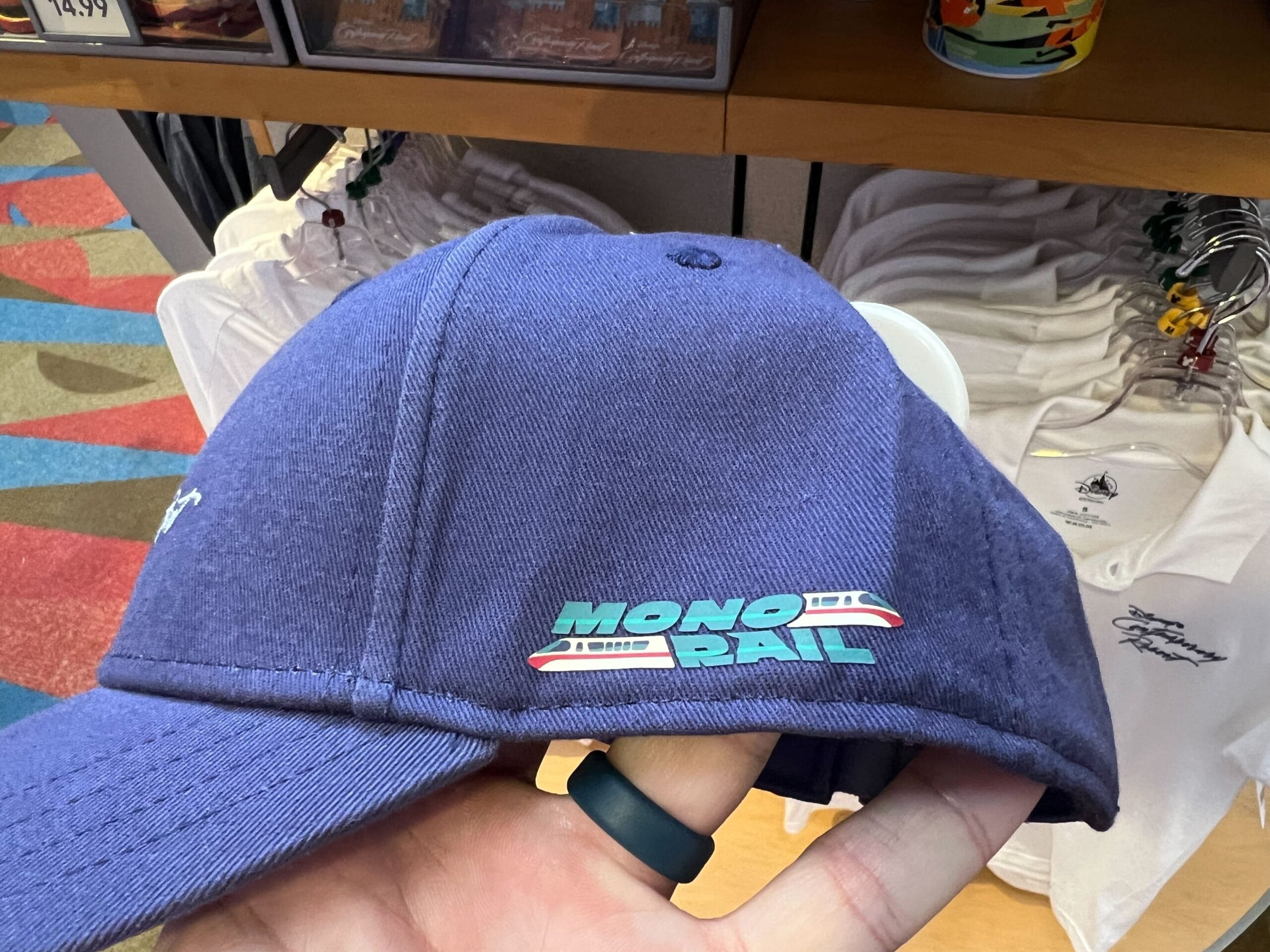 A hand holds a blue hat from the Monorail Collection, featuring Walt Disney World’s monorail and "MONORAIL" on the side.