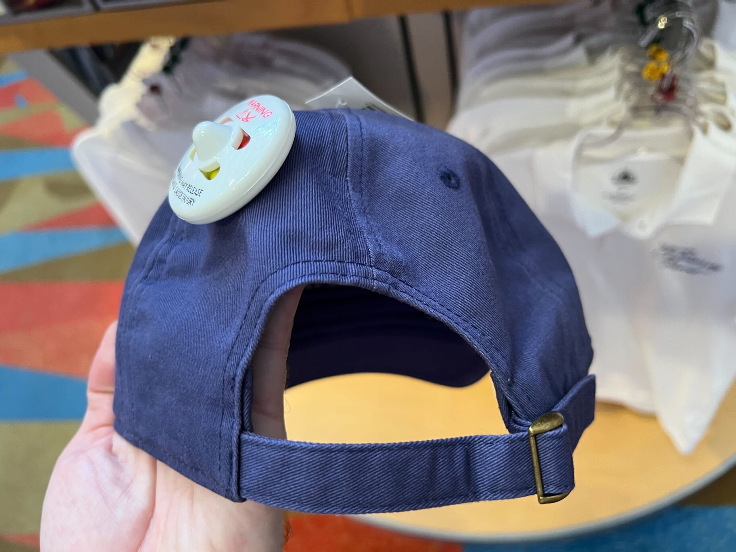 A hand holds a blue Monorail Collection cap with a security tag, displayed near white shirts at Contemporary Resort.