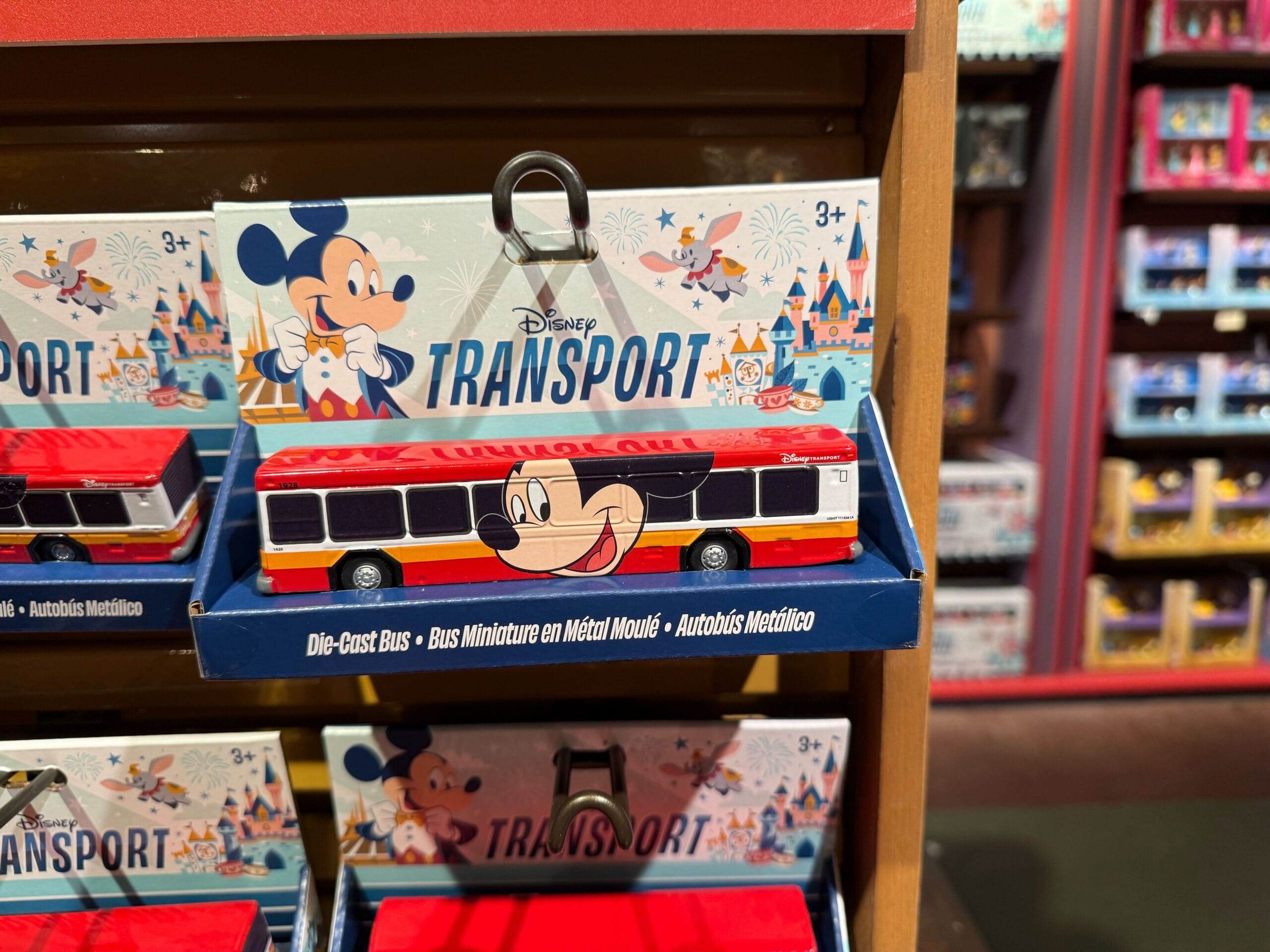 A toy Disney bus with Mickey Mouse design is displayed on a shelf in its packaging labeled “Disney Transport.”