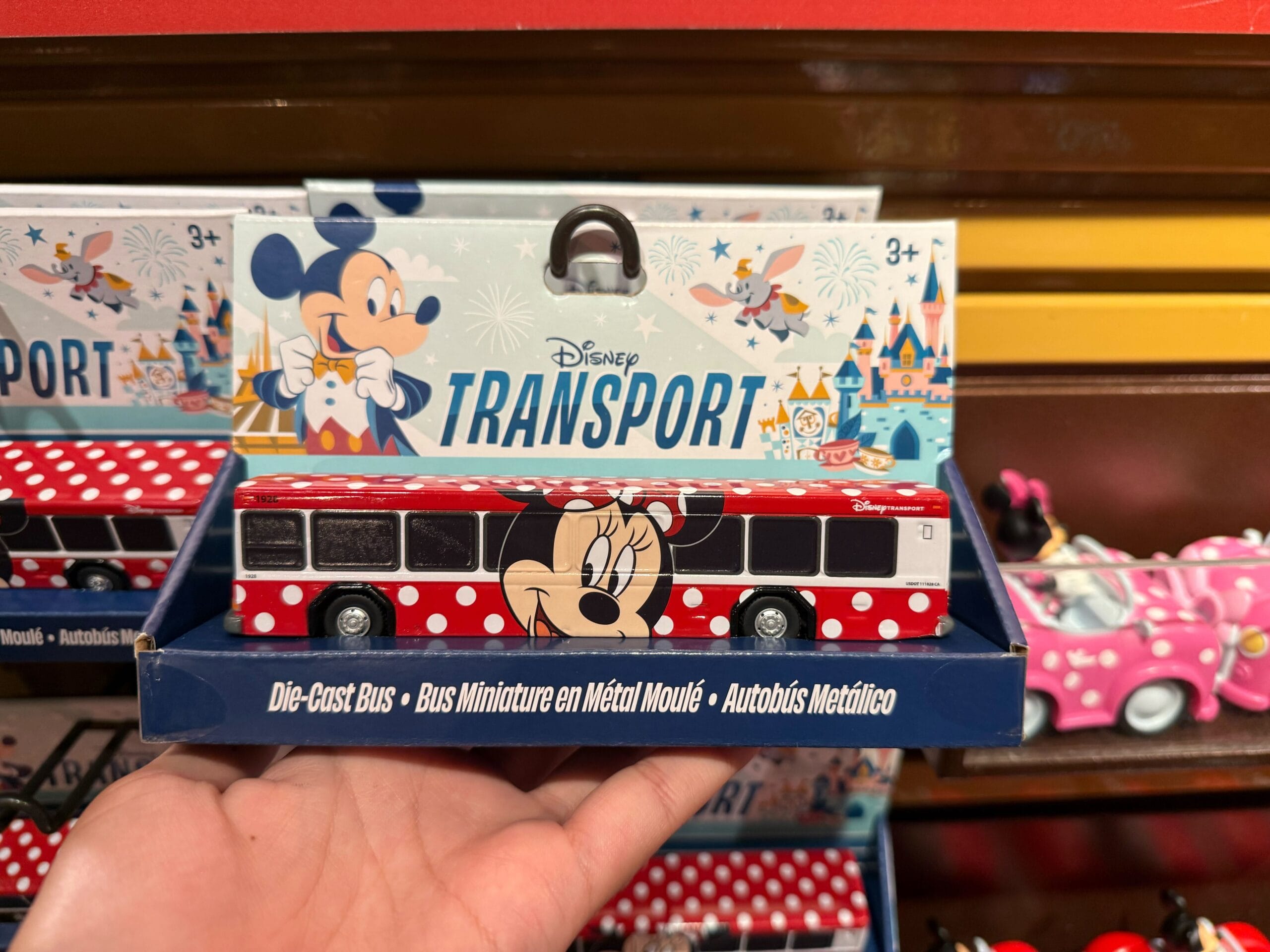 A hand holds a Disney Transport die cast bus featuring Minnie Mouse, with Mickey Mouse on the Disney Buses packaging behind.