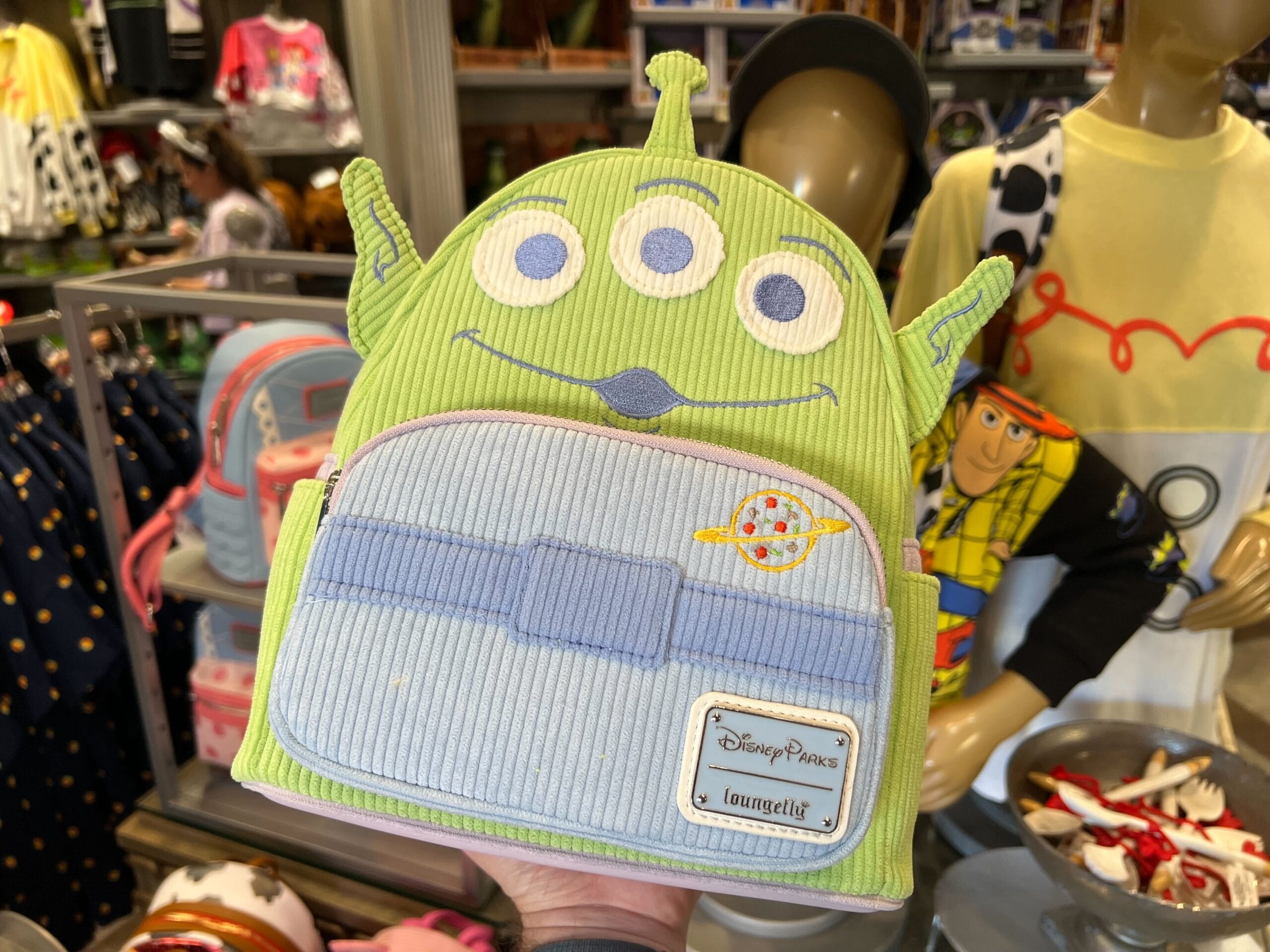 A hand holds a green Alien-themed Disney Parks Loungefly backpack in a store, with Toy Story merchandise in the background.