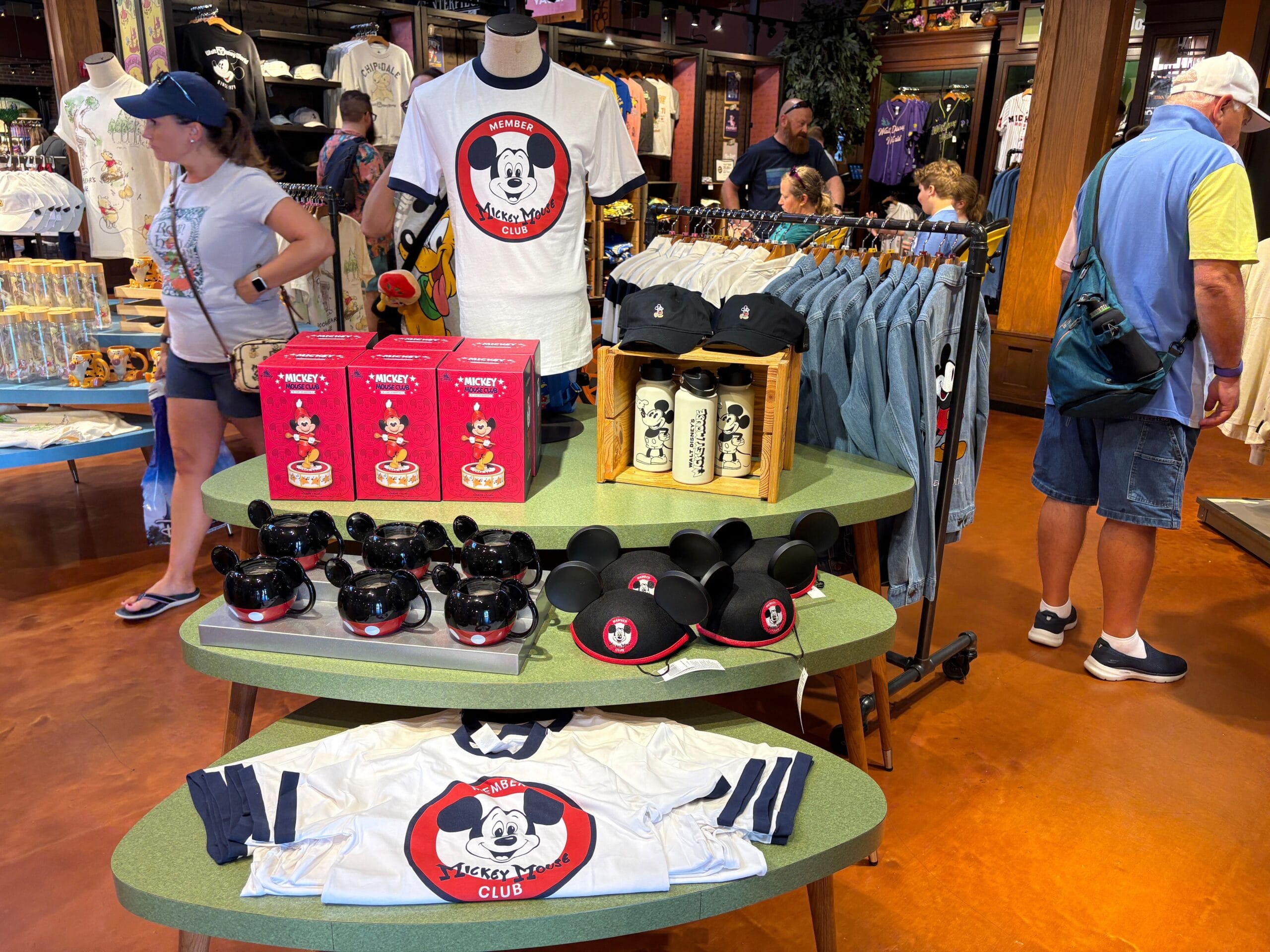 Display of Mickey Mouse Club 70th Collection merchandise in a store, with shirts, hats, water bottles, and people shopping.