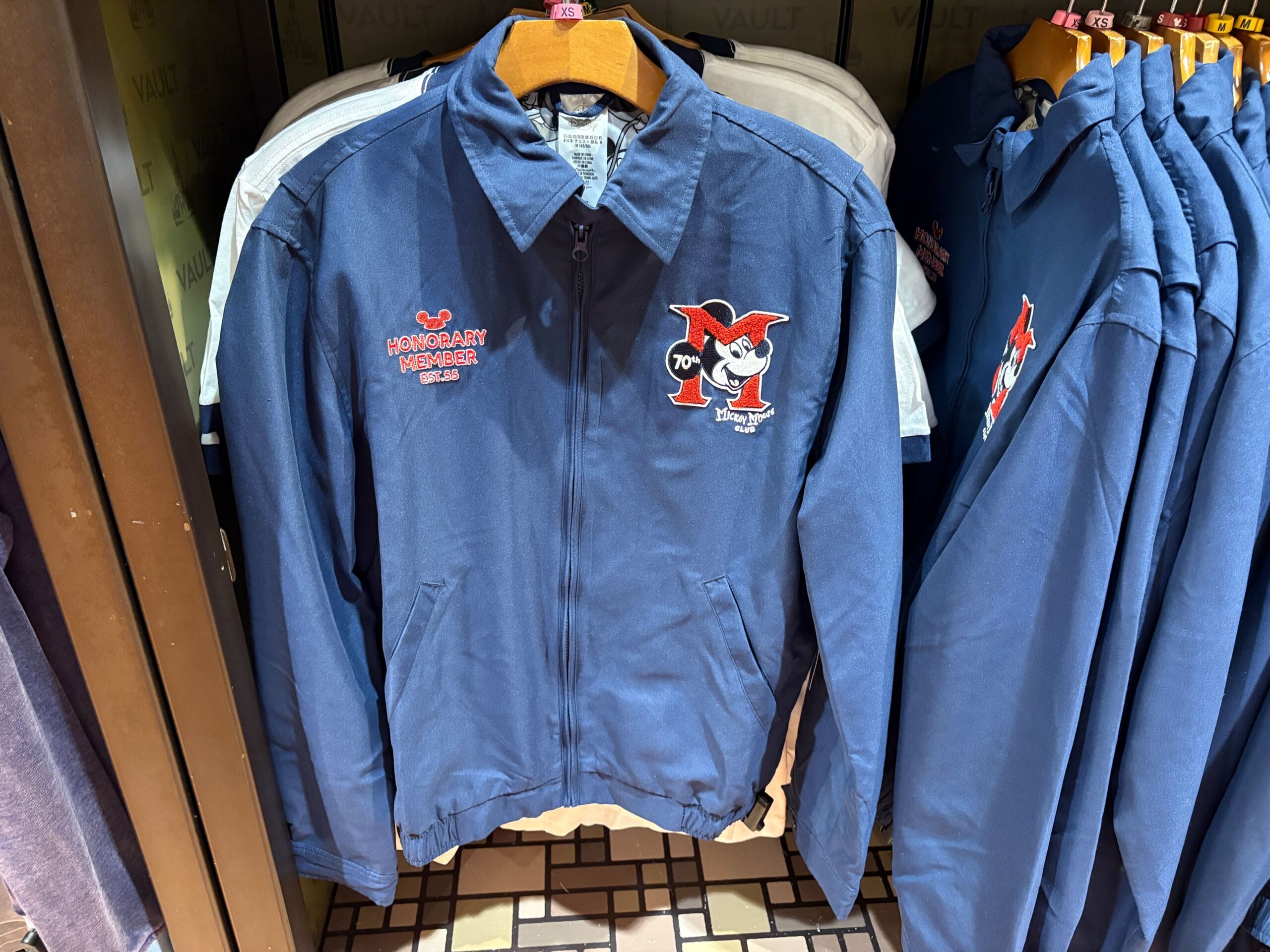 A blue jacket from the Mickey Mouse Club 70th Collection with "Honorary Member" text, hanging on a store rack.