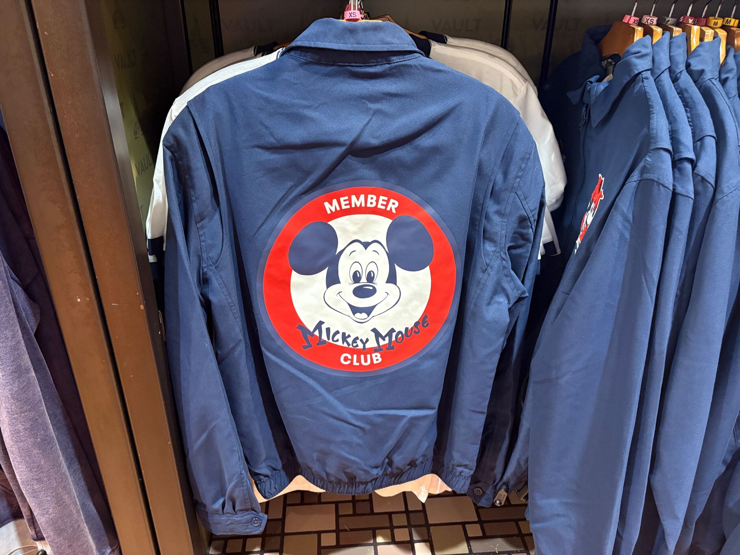 Blue jacket from the Mickey Mouse Club 70th Collection, featuring a large Mickey face, hanging on a rack.