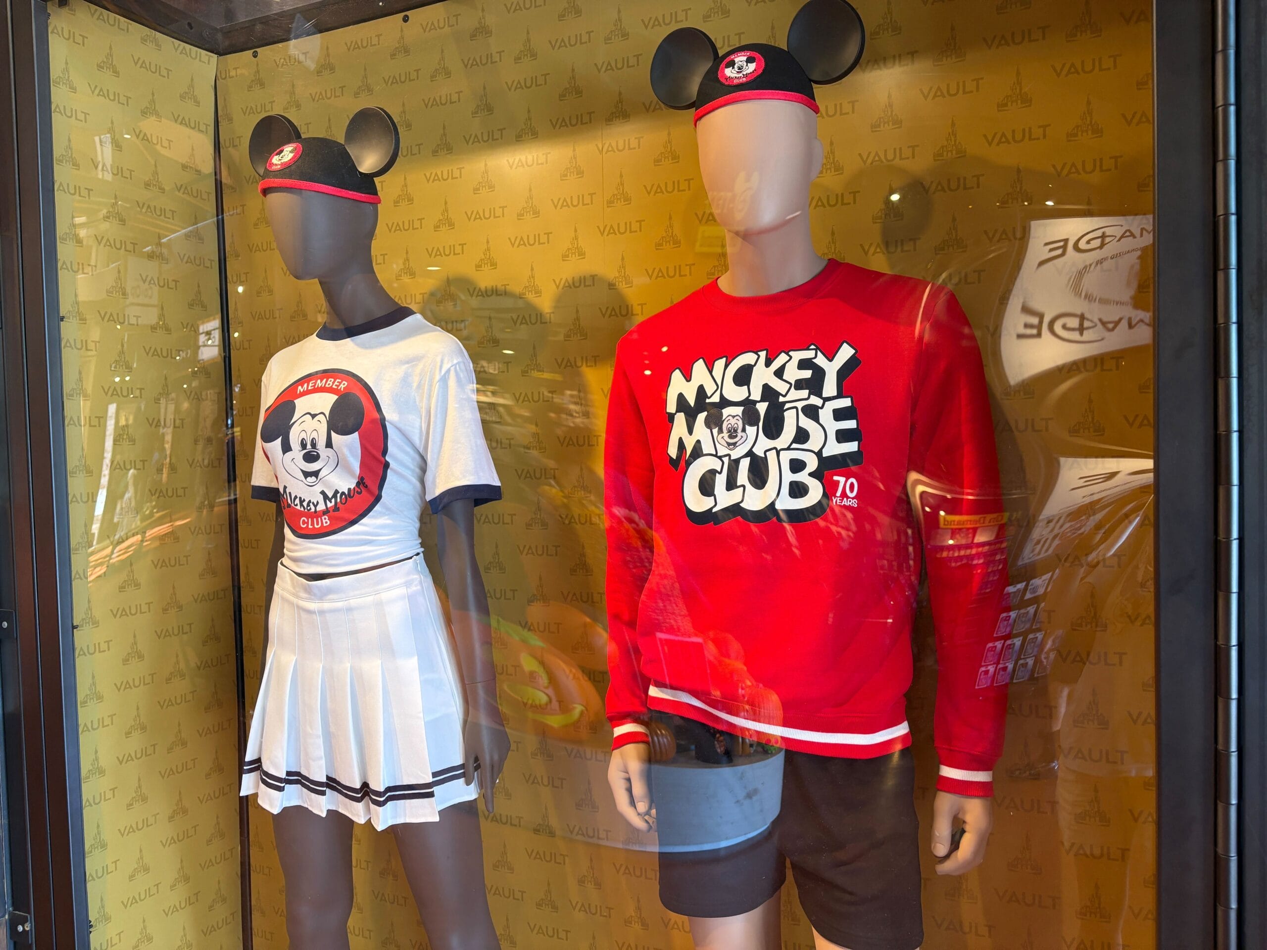 Two mannequins wear Mickey Mouse Club outfits and ear hats in a display case with a yellow VAULT 70th Collection background.
