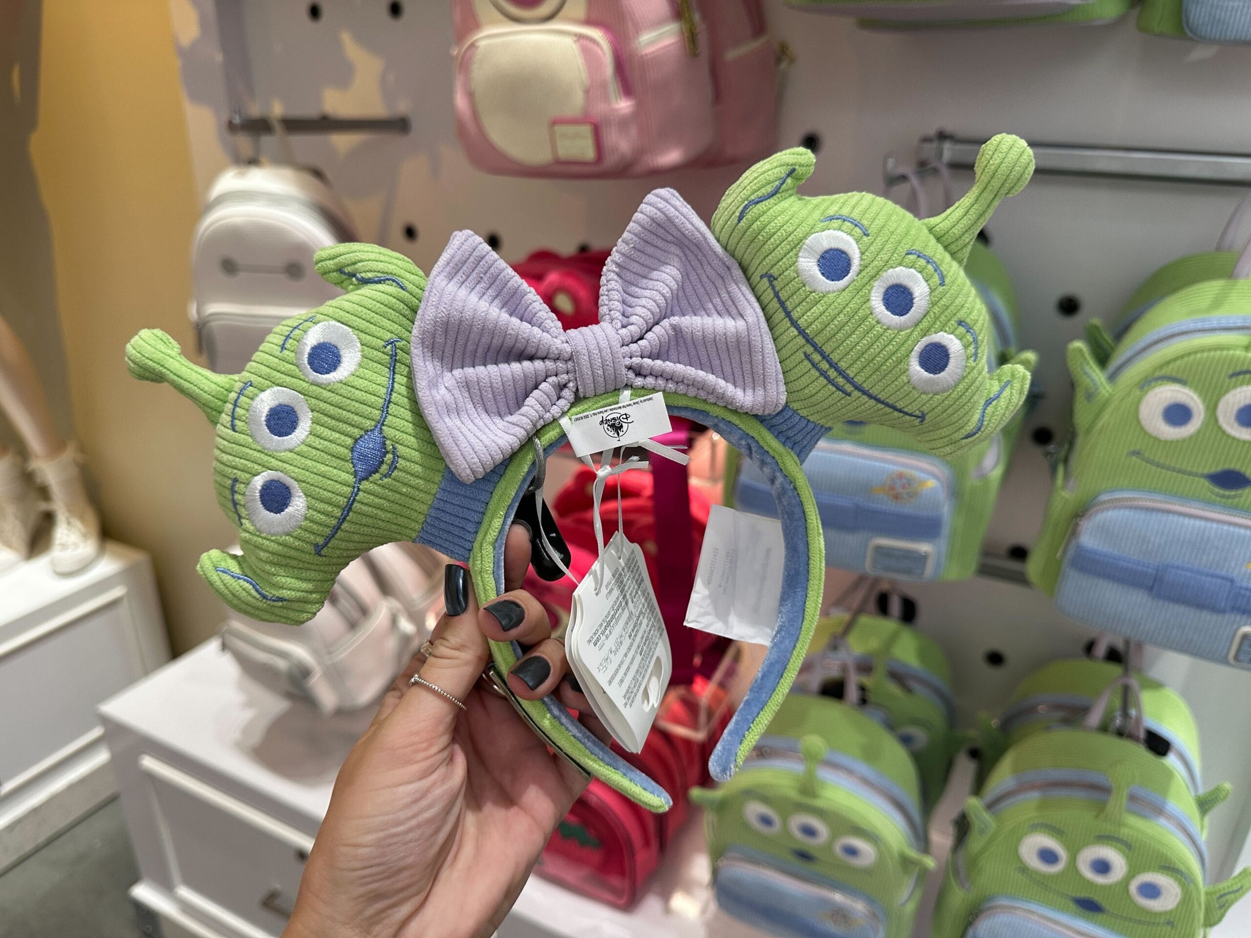 A hand holds a Toy Story Alien headband with a purple bow, displayed in a store featuring the Loungefly Collection.