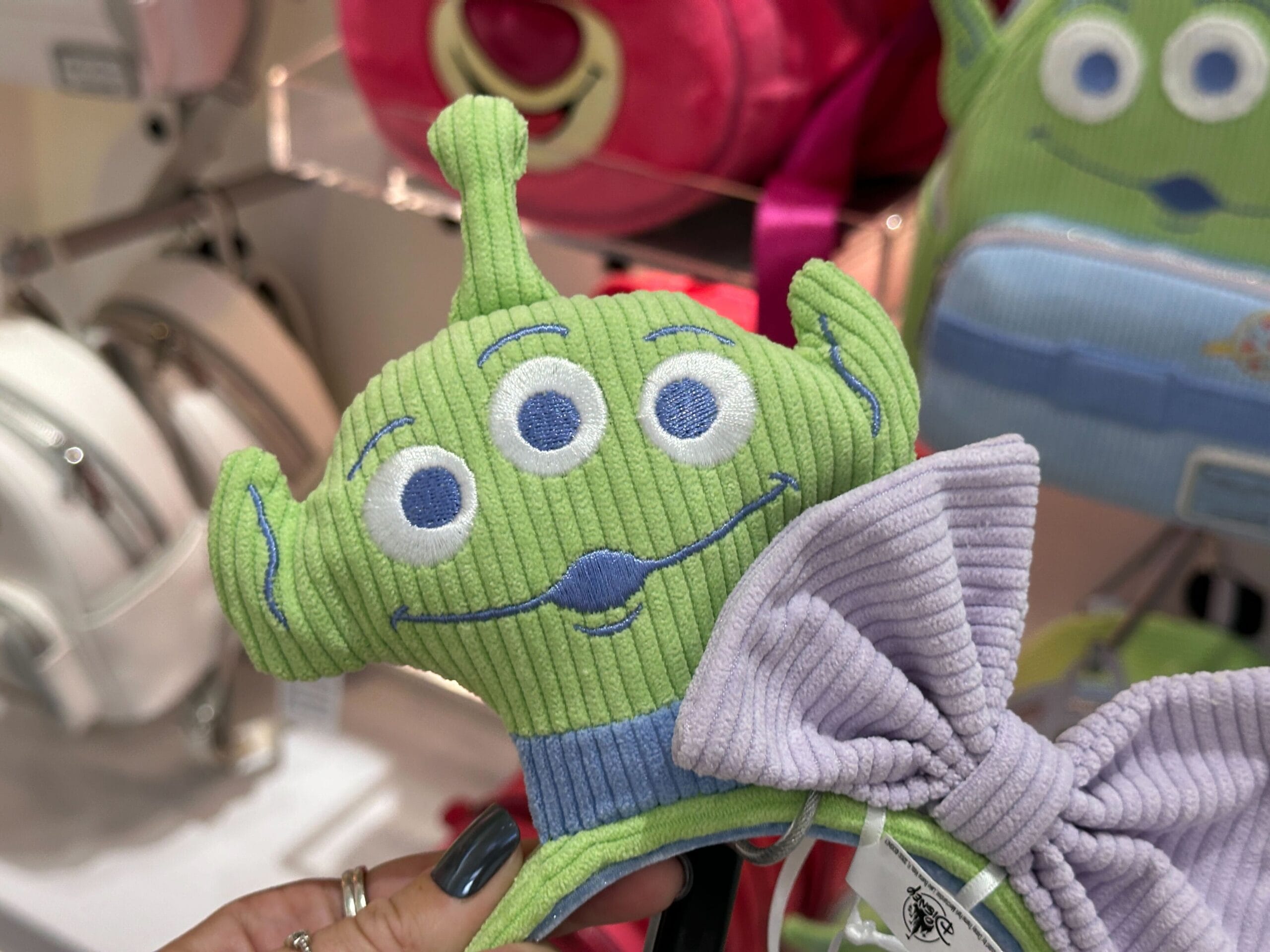A plush green Toy Story alien head with three eyes and a big lavender bow, held up in a store display.