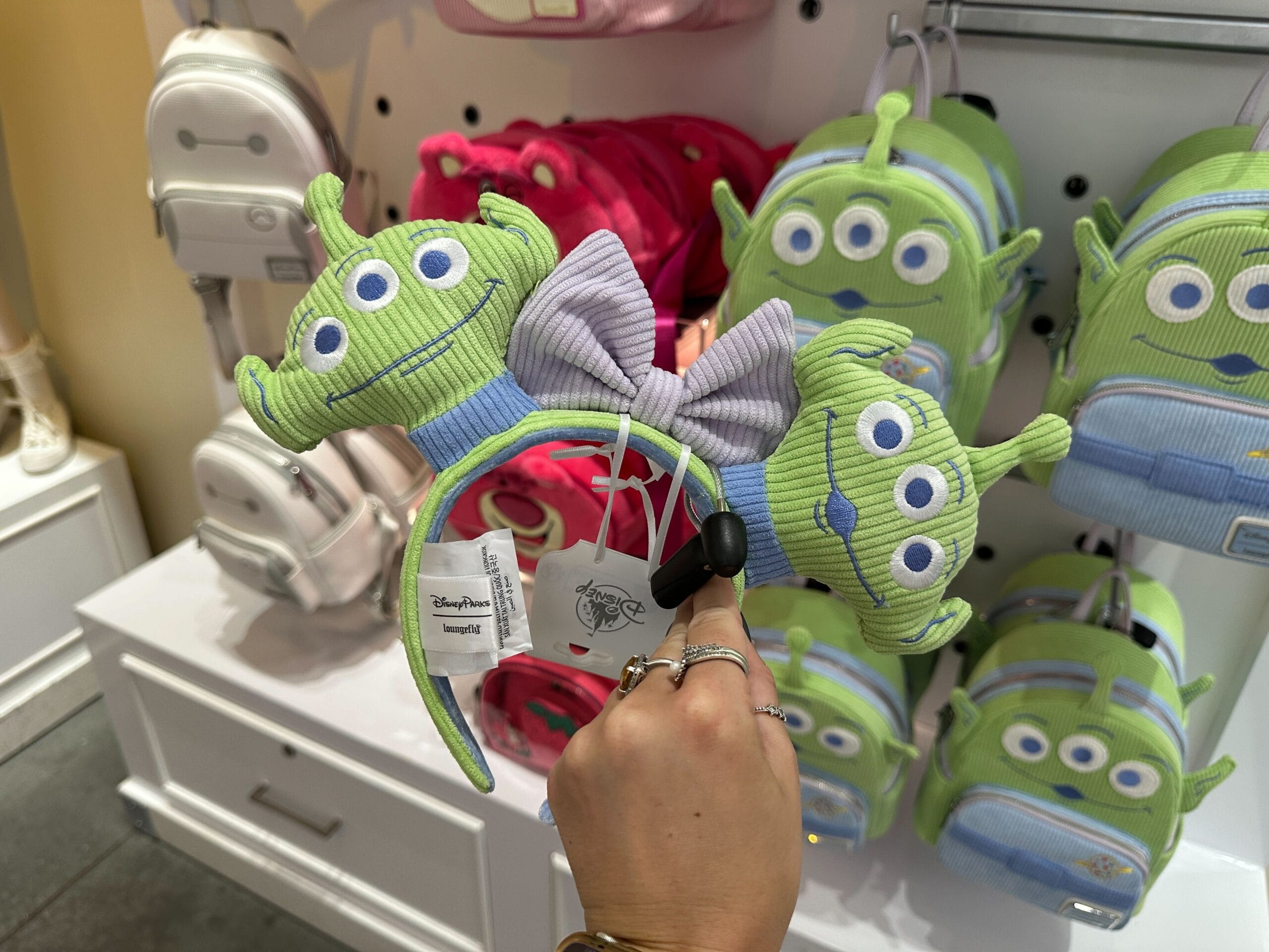 A hand holds a Toy Story alien headband with a bow, surrounded by pastel Toy Story Alien Loungefly backpacks in a store.