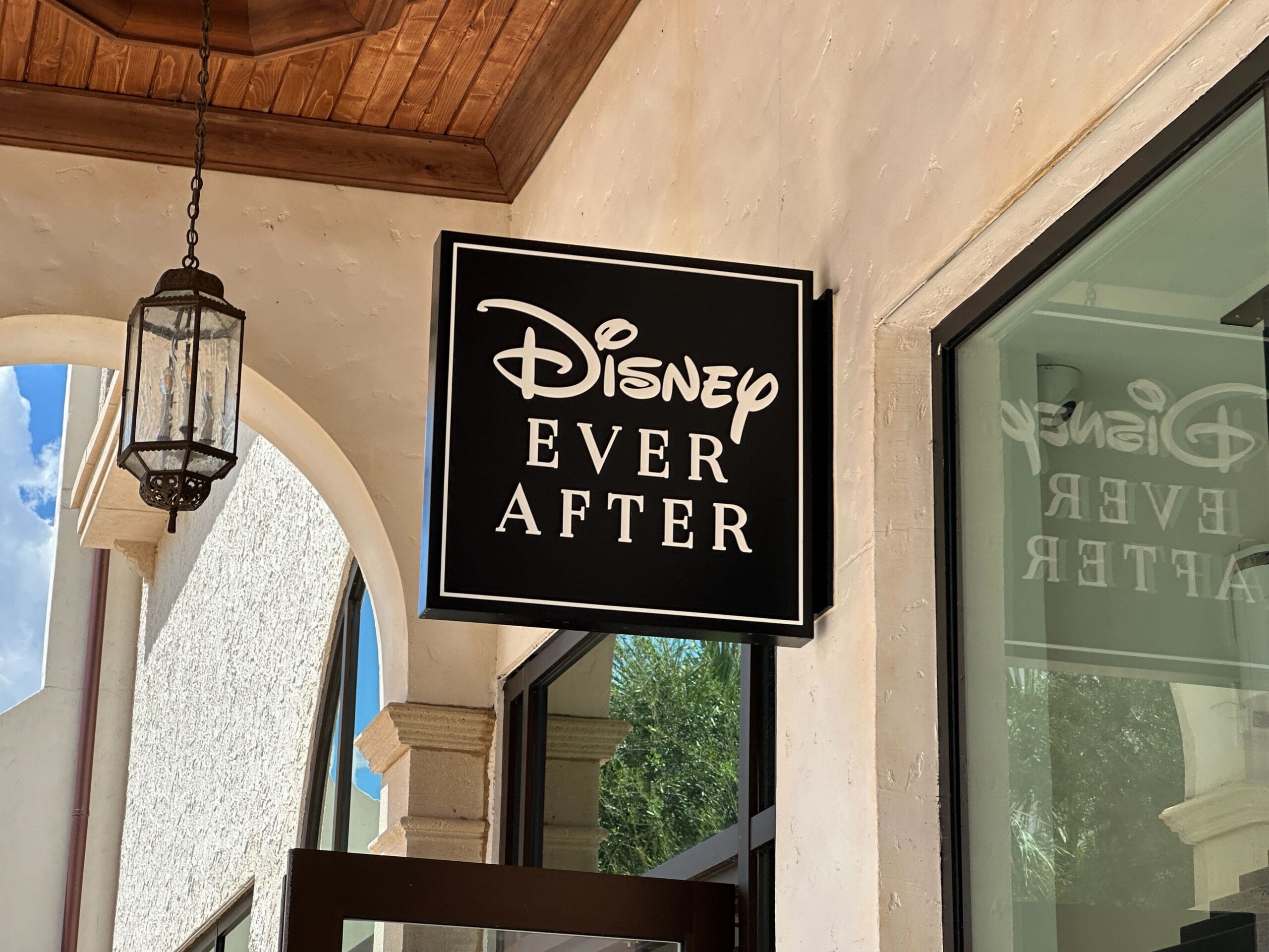 A black and white sign reading "Disney Ever After" hangs outside a building at Disney World with arched windows and a lantern.