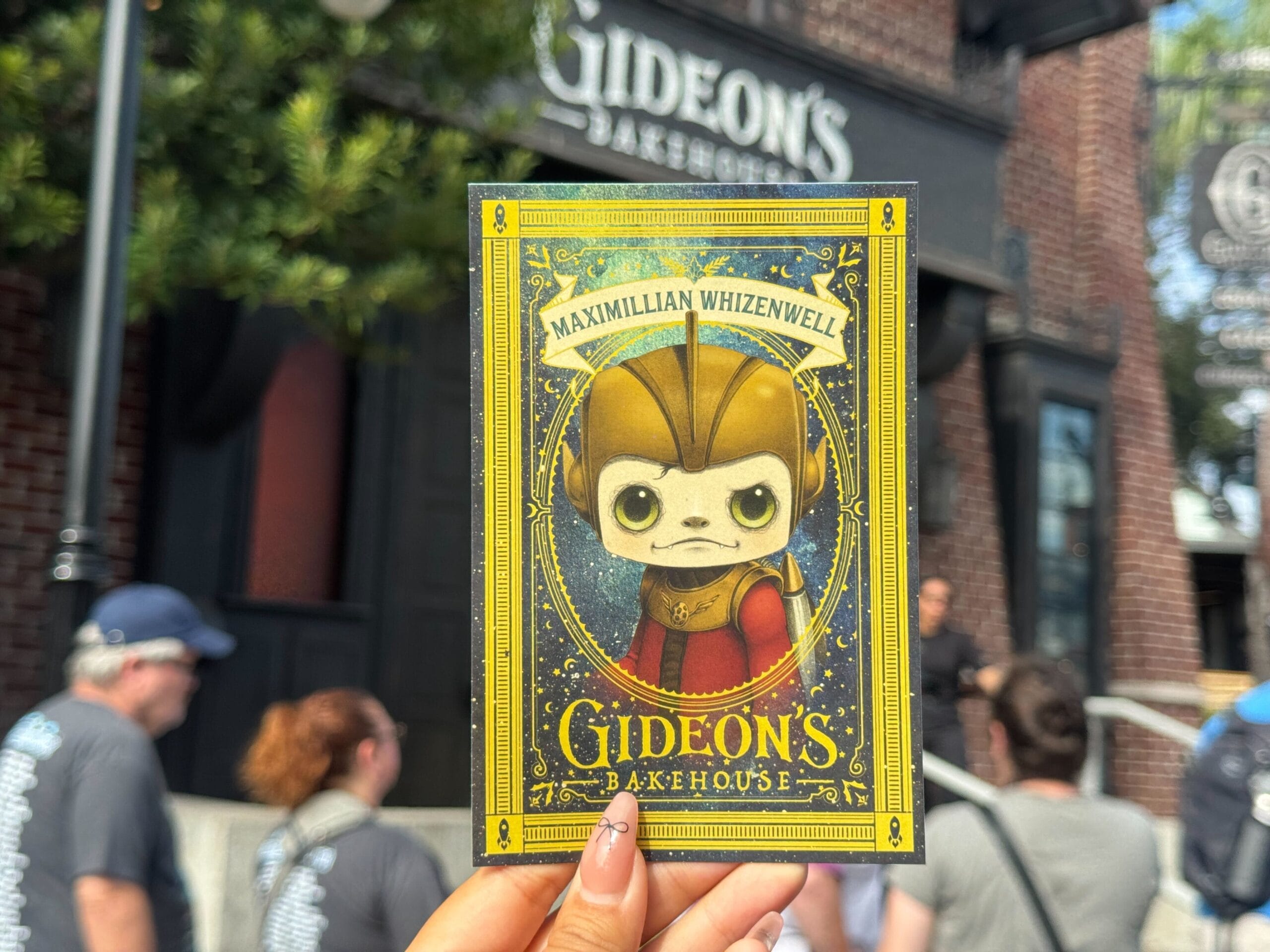 A hand holds a Gideon's Bakehouse card featuring a character named Maximilian Whizenwell outside the store.