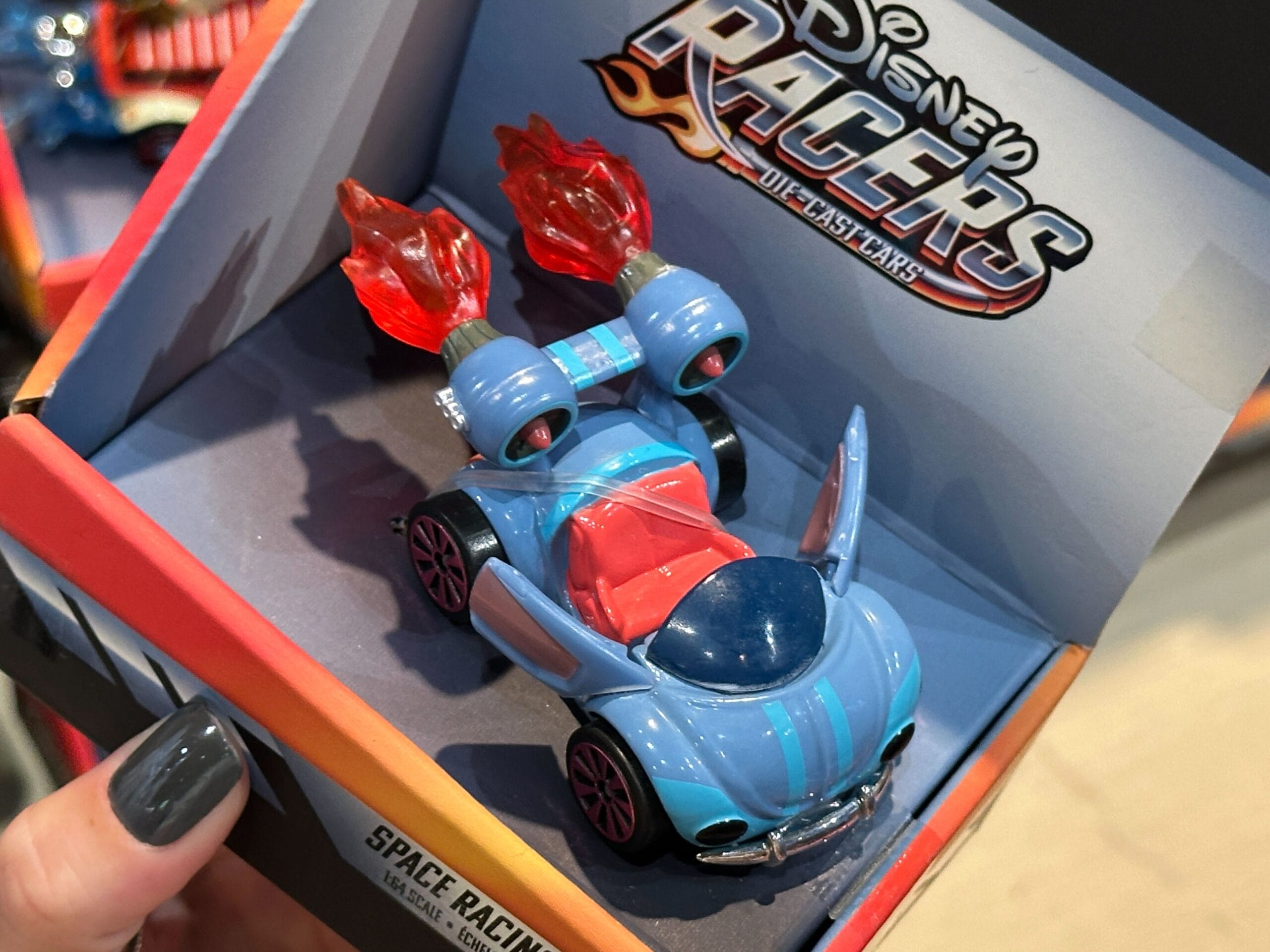 A blue Disney Racers toy car with red flames, in packaging, held by a hand with gray painted nails.