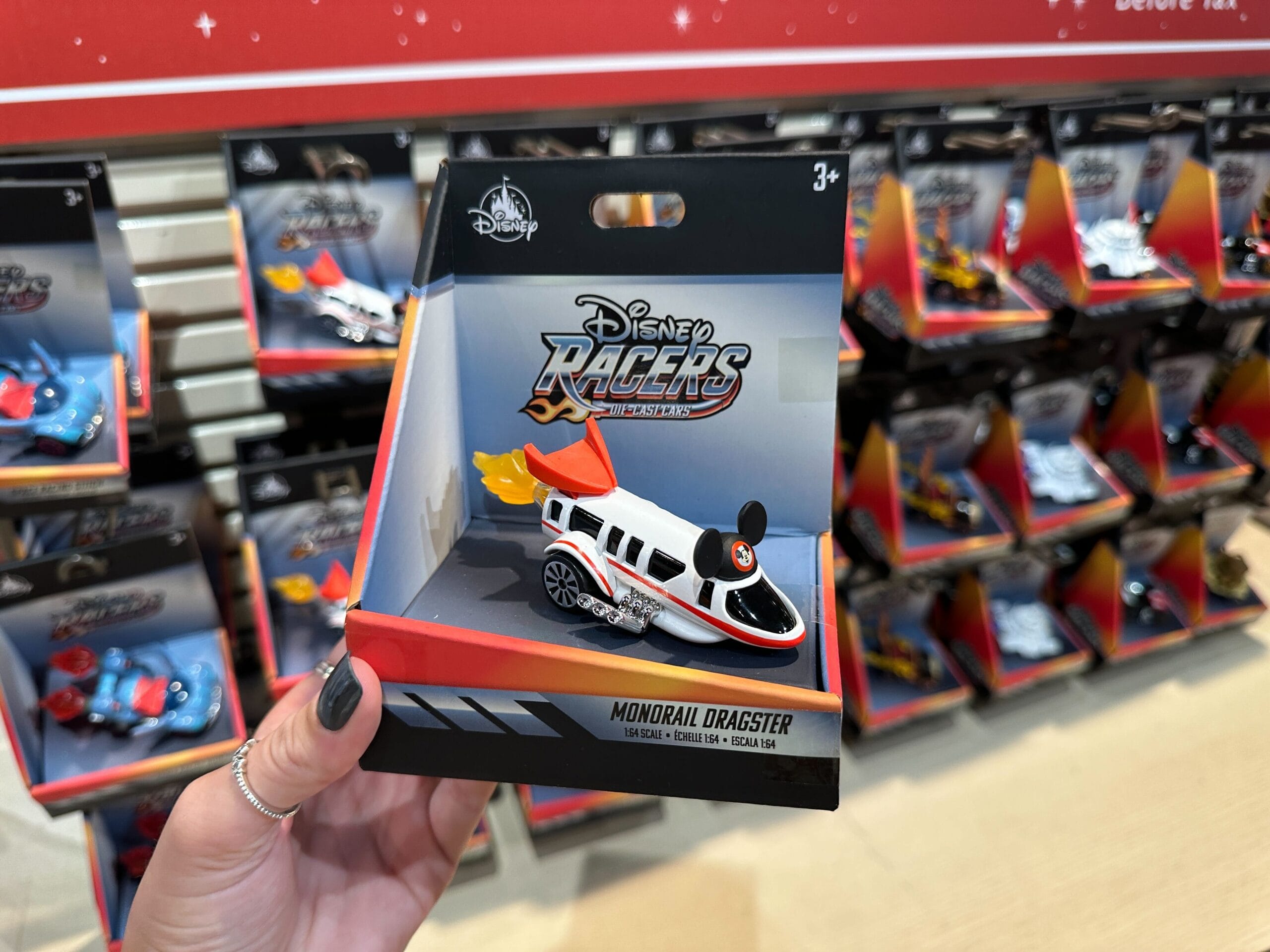 A hand holds a Disney World Racers Monorail Dragster toy car in a store display with more toy cars in the background.