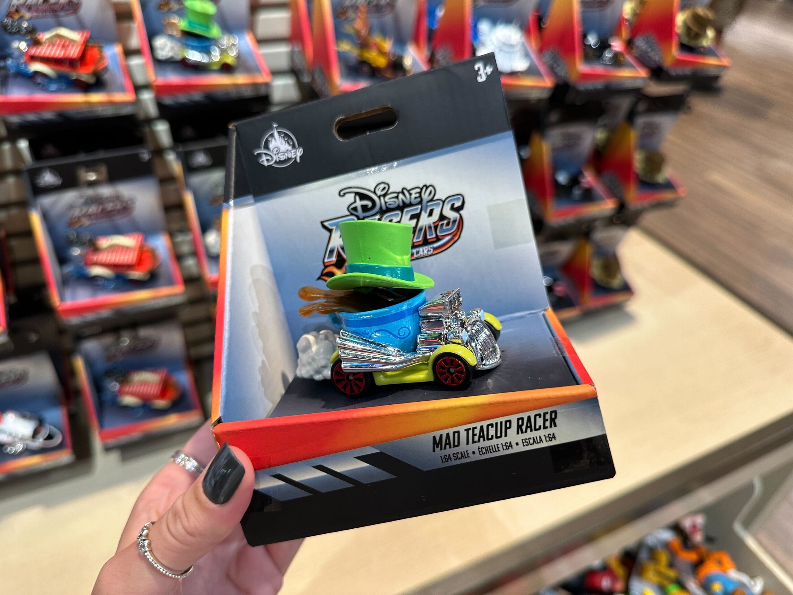 A hand holds a Disney Racers Toy "Mad Teacup Racer" in a store at Disney World, with more toys on shelves behind.