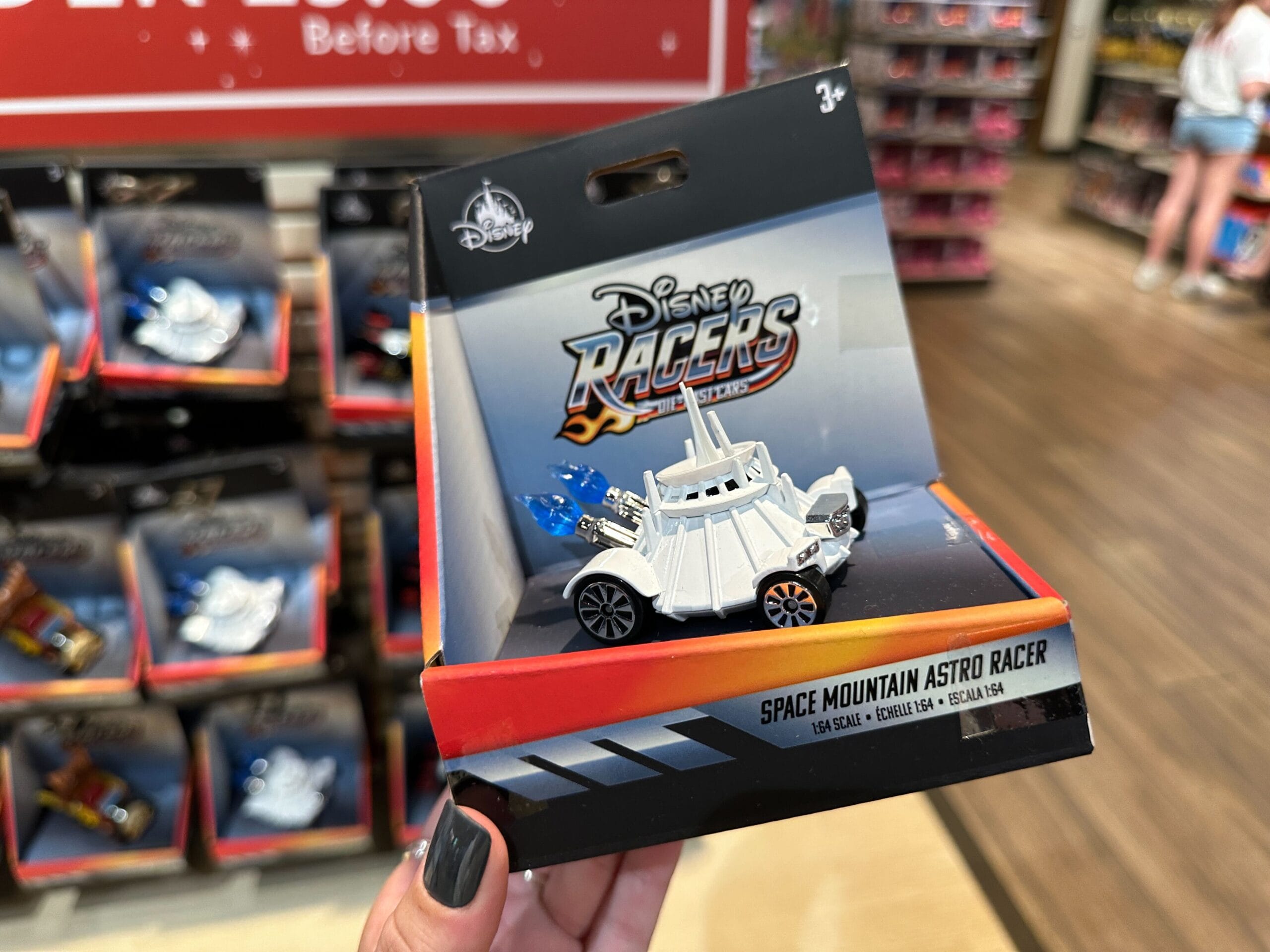 A hand holds a Disney Racers Space Mountain Astro Racer toy car in a Disney World store aisle with toys on shelves.