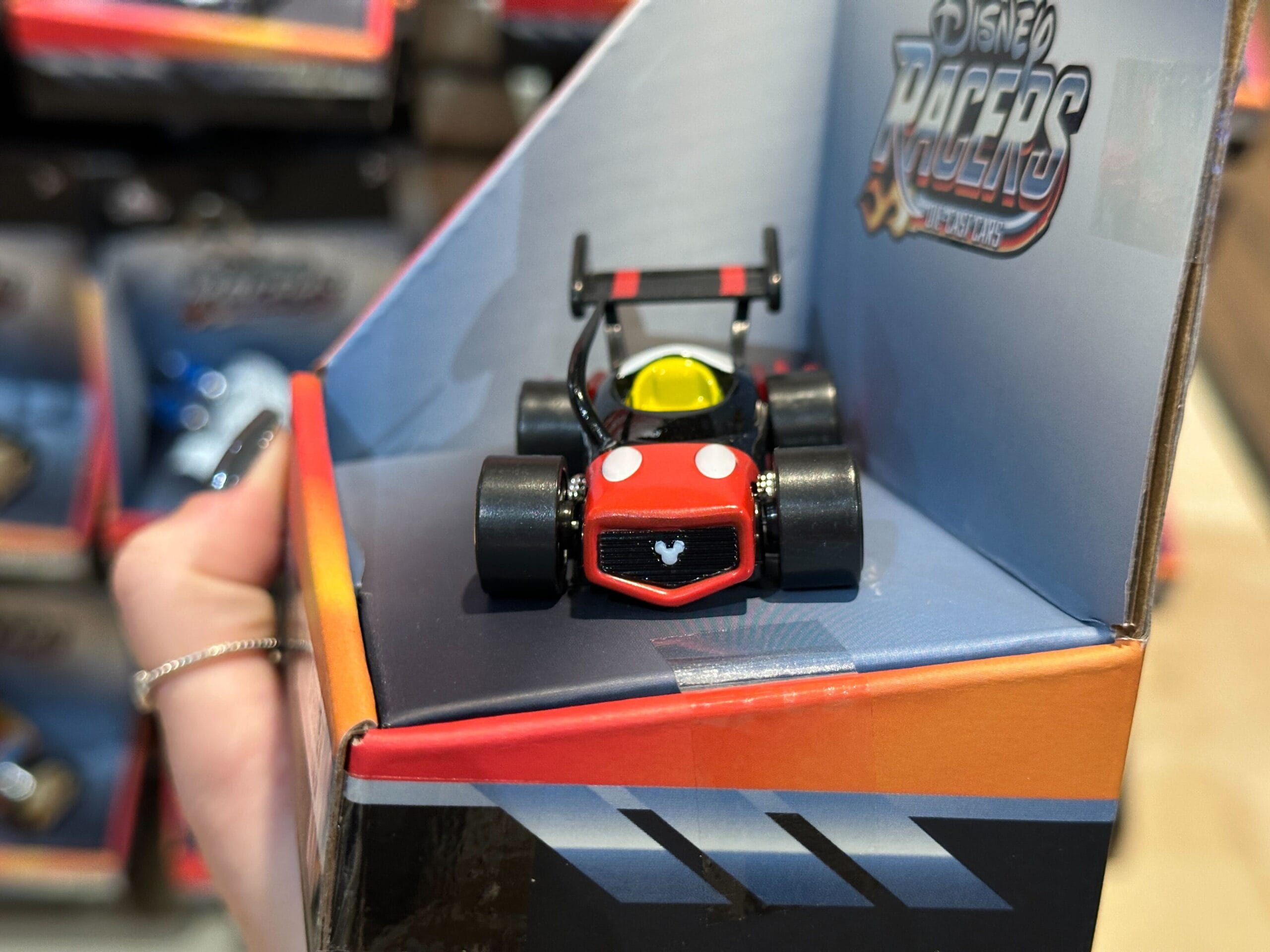 A hand holds a Disney Racers toy car with Mickey Mouse details, inspired by the Jungle Cruise, in a store display box.