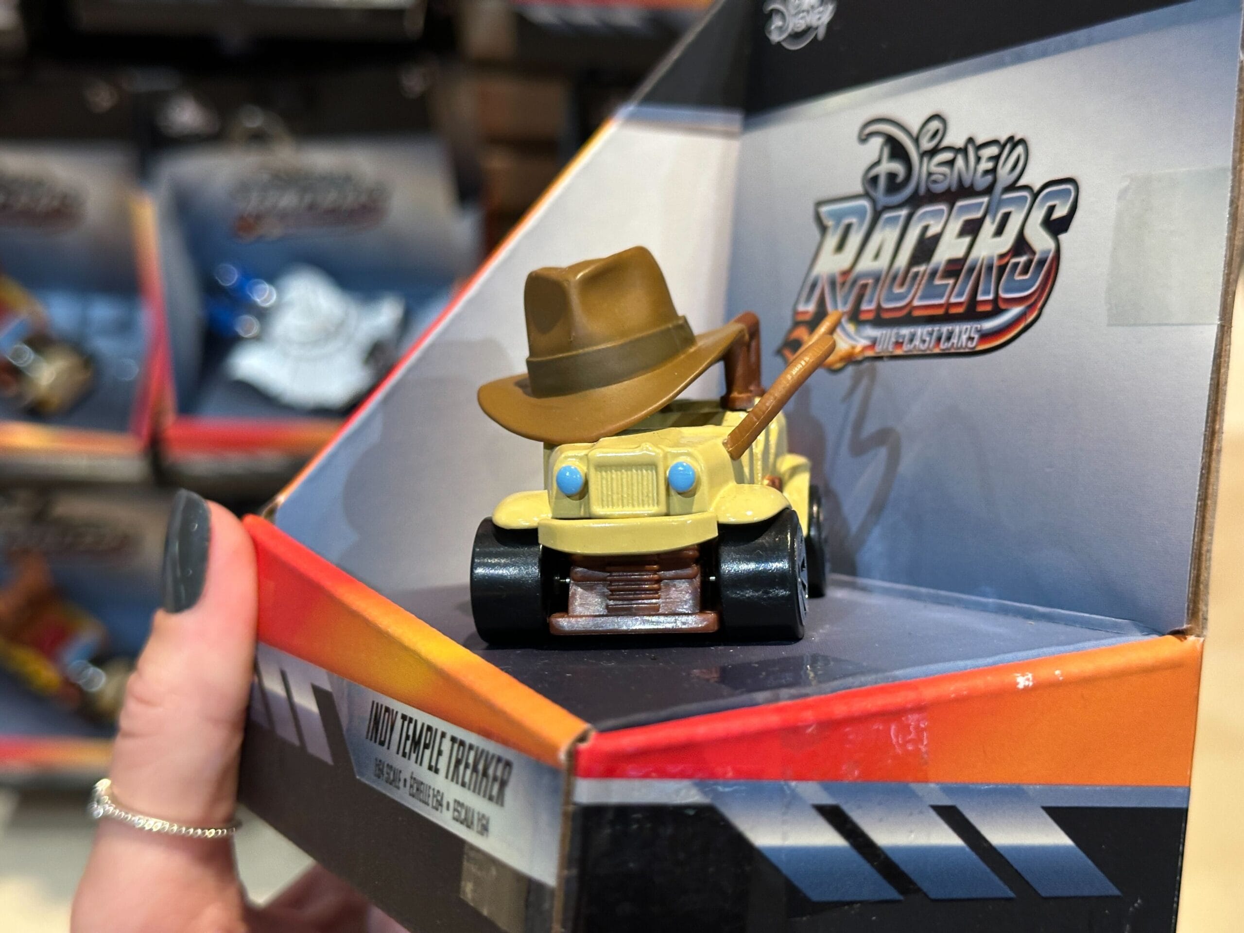 A hand holds a Jungle Cruise toy car with a brown hat and whip, displayed in Disney Racers packaging.