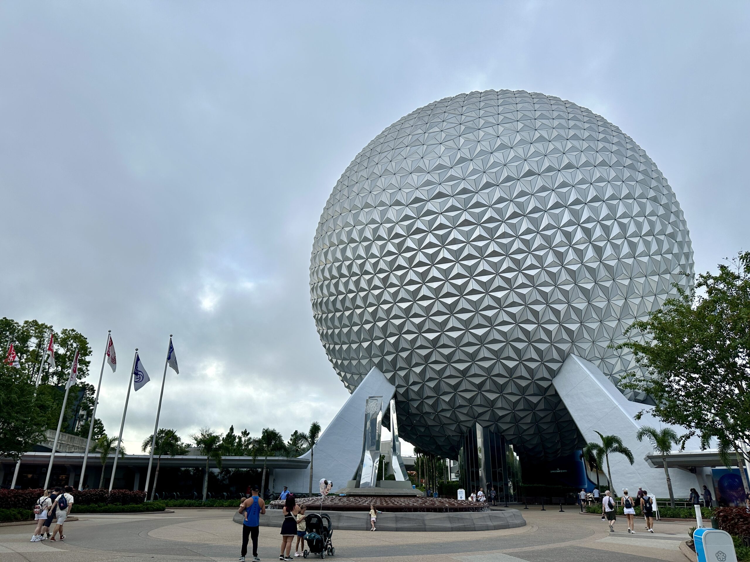 A large geodesic sphere stands at EPCOT with people and flags in front, celebrating Food and Wine under a cloudy sky.