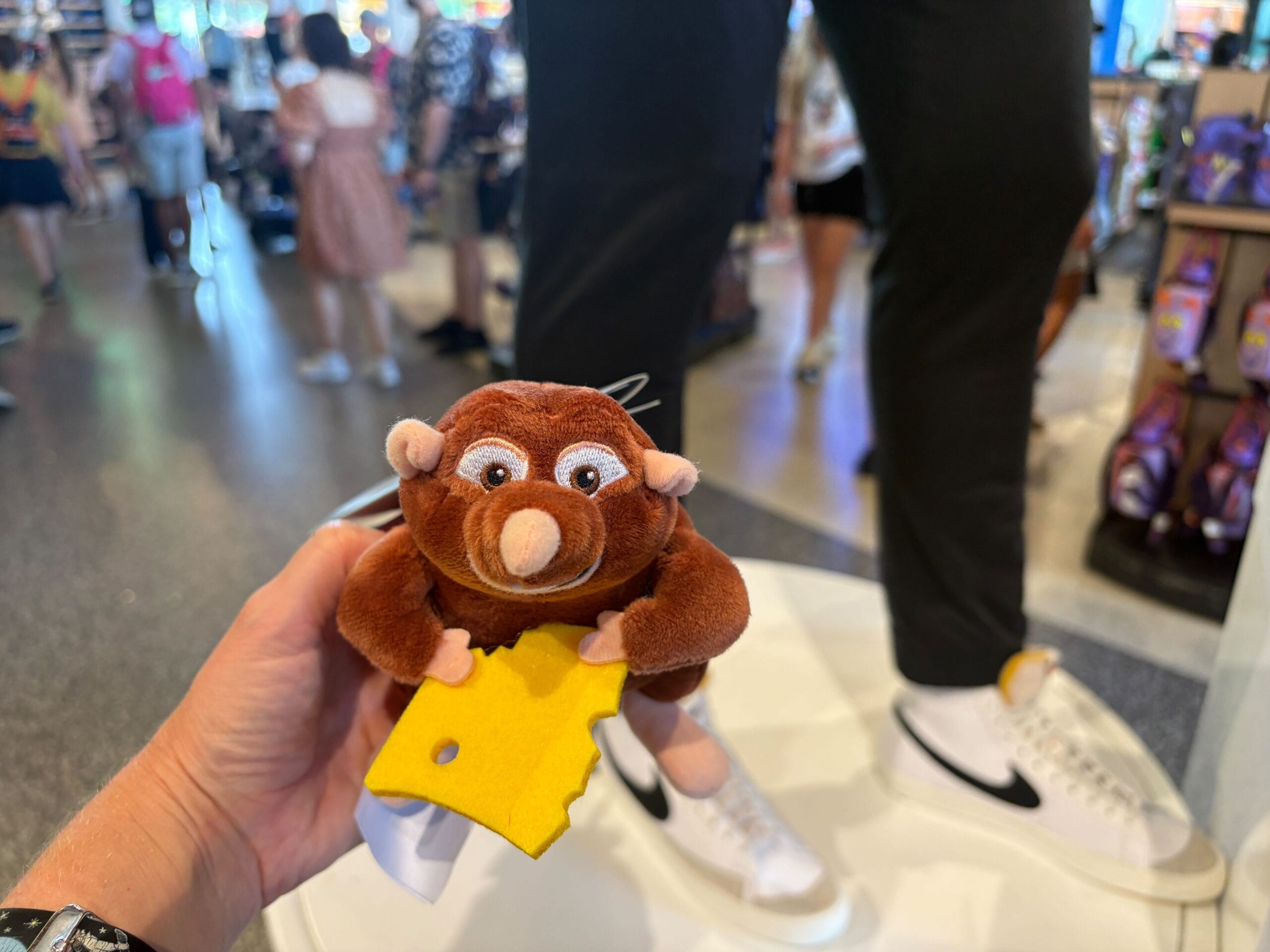 A hand holds a brown plush rat with a yellow cheese slice inside a busy store with people and displays in the background.