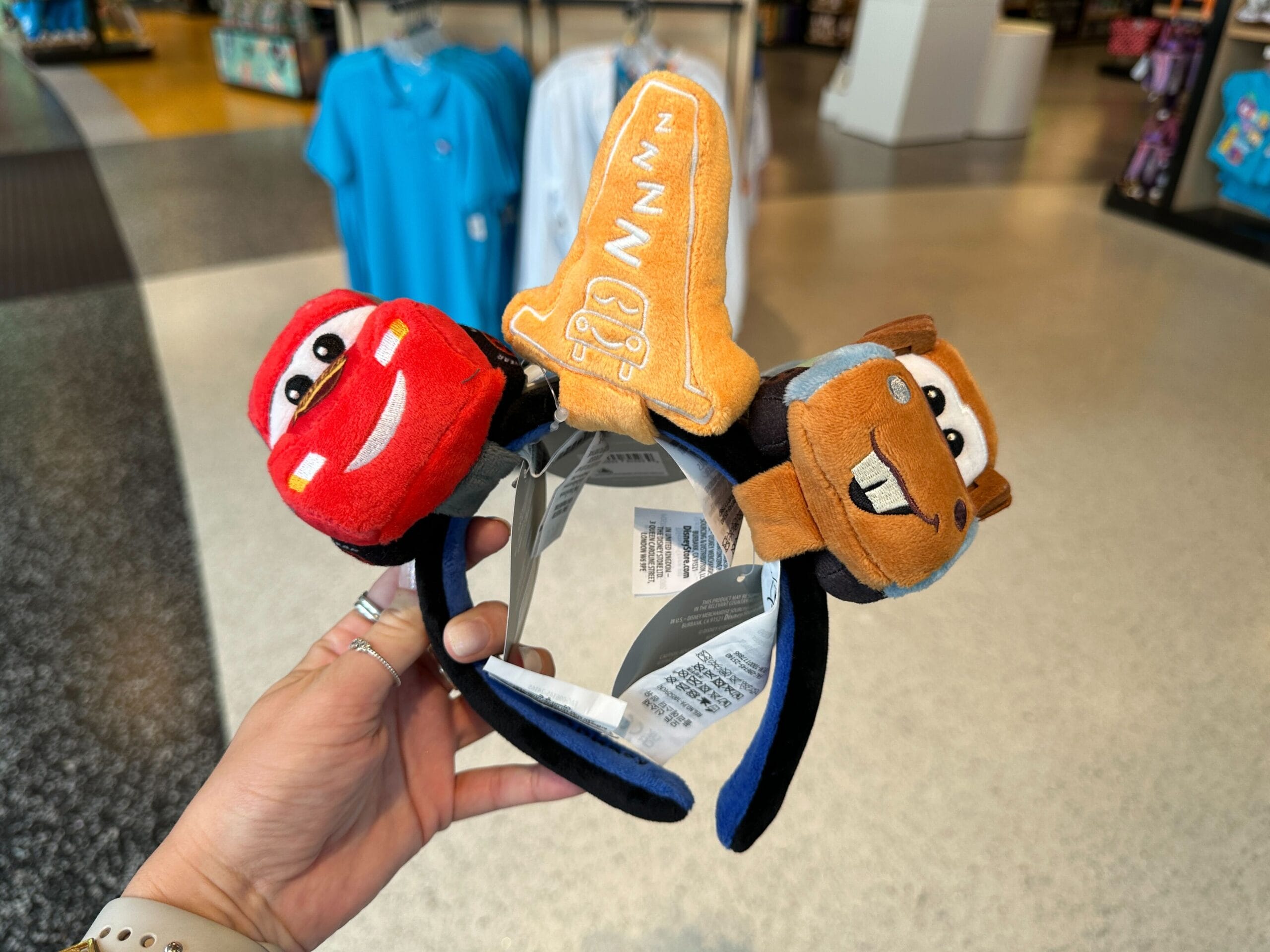 A hand holds a Cars Plush Headband with Lightning McQueen, Mater, and a “1” pennant—perfect for Disney World fans.