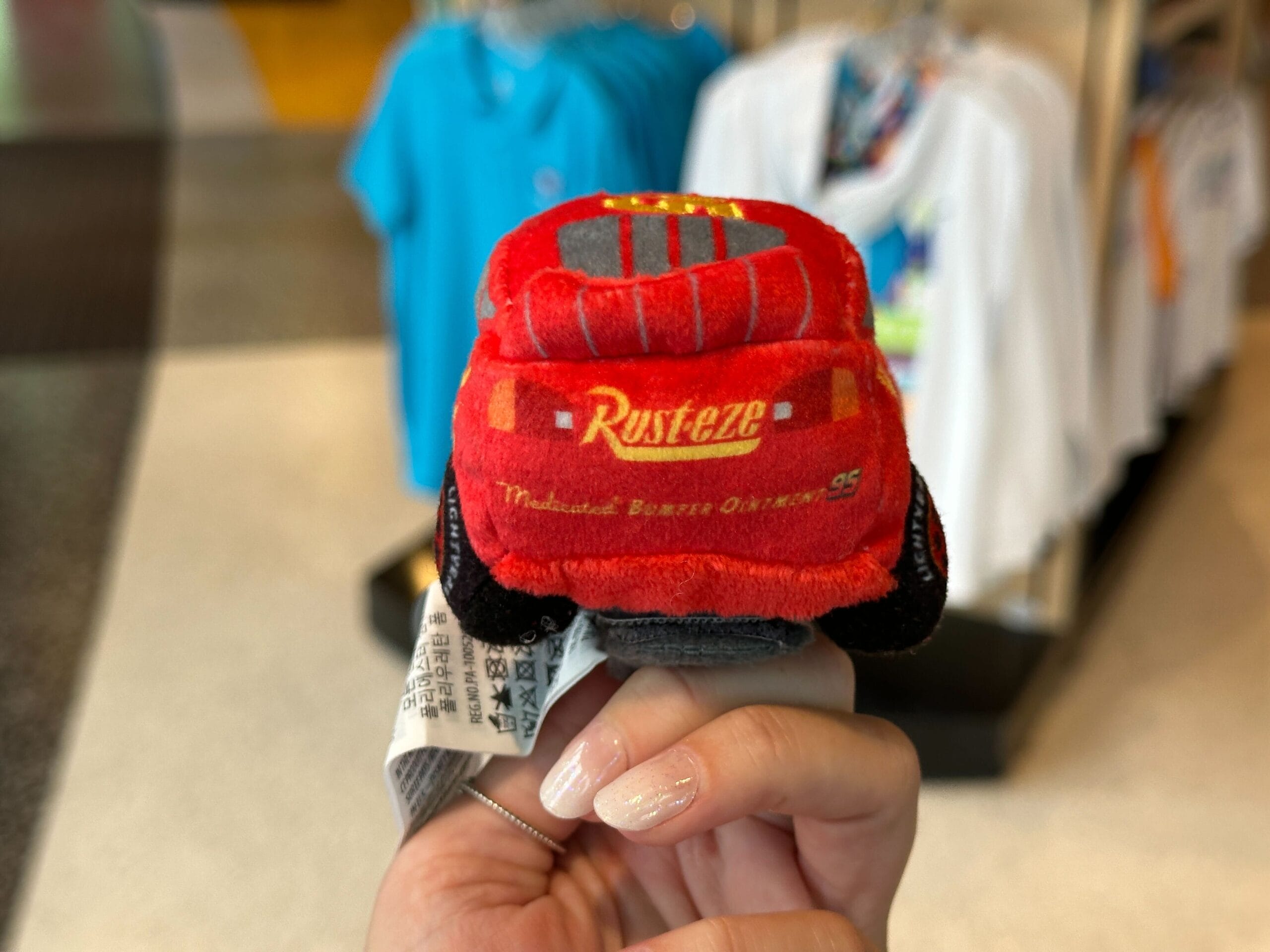 A hand holds a Lightning McQueen plush toy at Disney World, with shirts on racks in the background.
