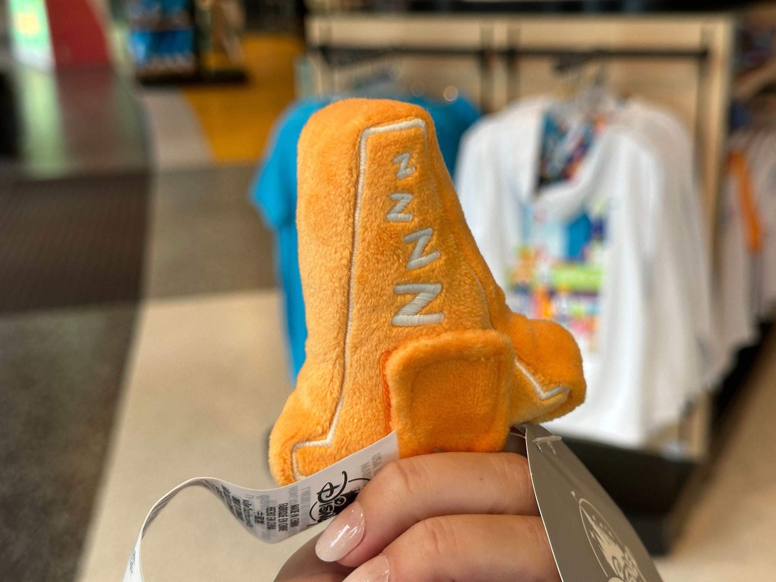 A hand holds an orange rocket plush with "ZZZ" stitched on the side; Disney World shirts hang in the background.