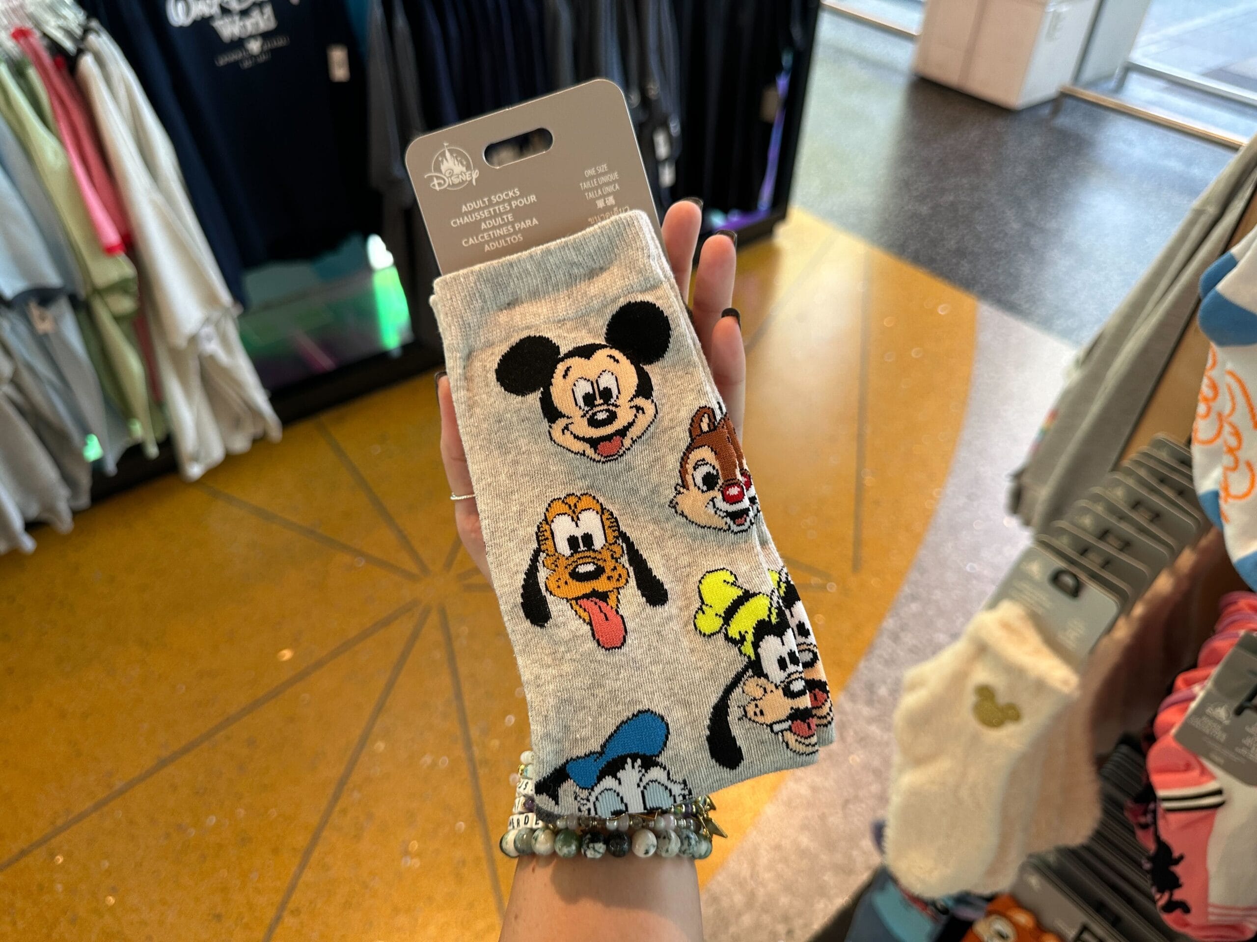 Hand holding Disney World novelty socks featuring Mickey, Pluto, Goofy, Donald, and Chip 'n' Dale in a store.