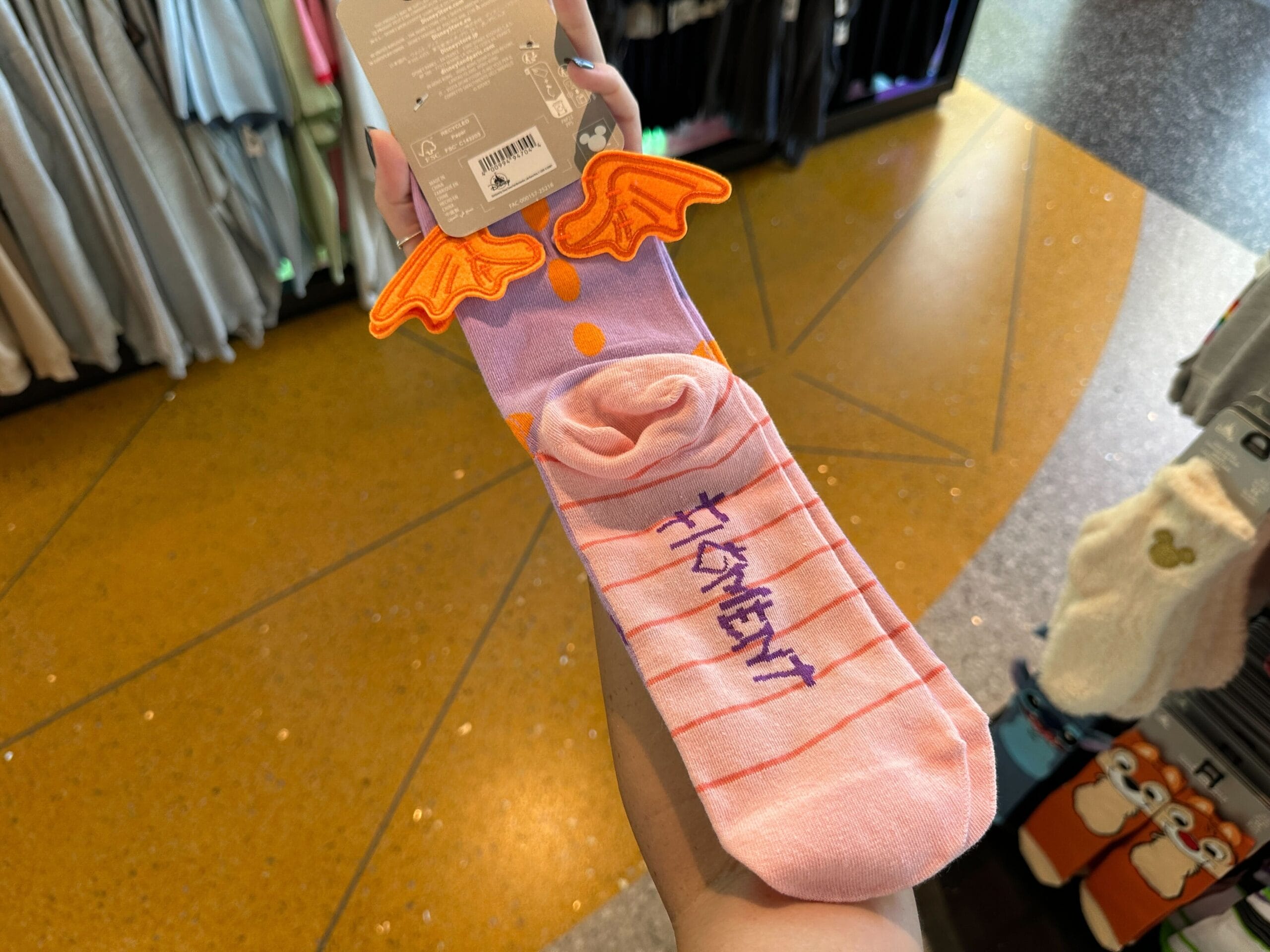 A hand holds Disney World Figment socks—novelty purple and pink with orange wings and "FIGMENT" in blue on the foot.