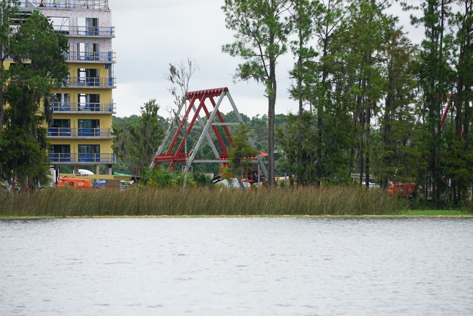 Lakeshore Update: A construction site with a yellow building and red steel structures near a lake, surrounded by tall trees.