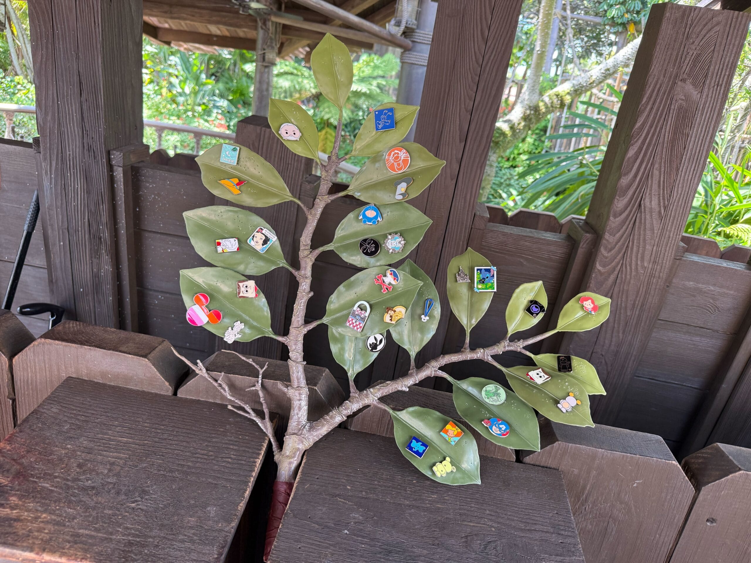 A branch with green leaves, each decorated with colorful pins, set against a wooden fence background.