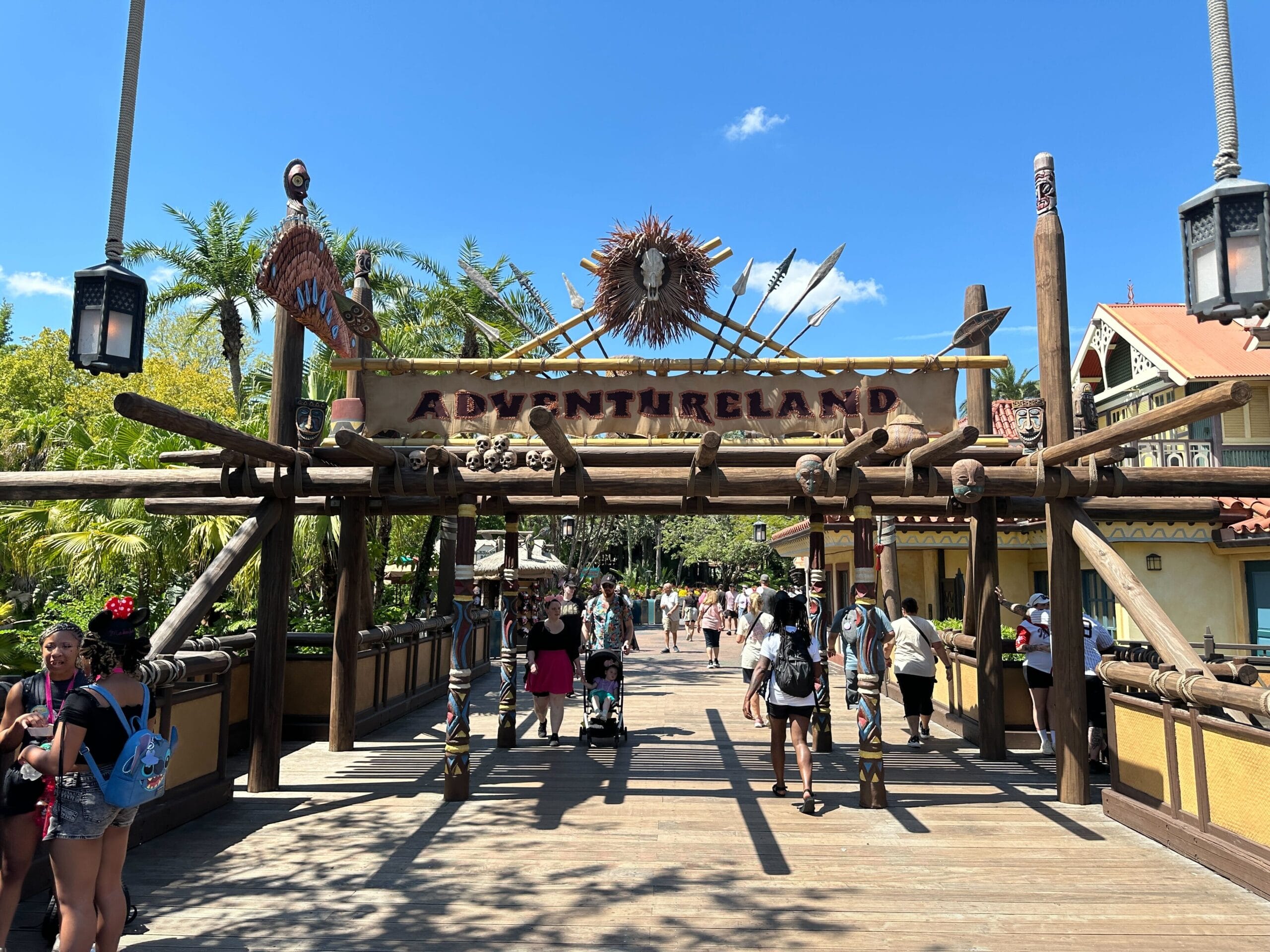 Guests stroll under the Adventureland Sign at Magic Kingdom, passing tribal designs—but notice some missing props above.