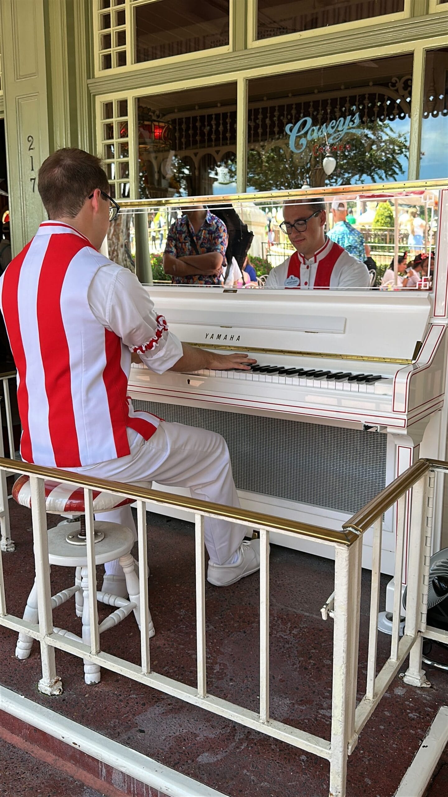 Grayson Smith, a Casey's Corner Pianist, plays a white piano as his image reflects in the mirror above the keyboard.