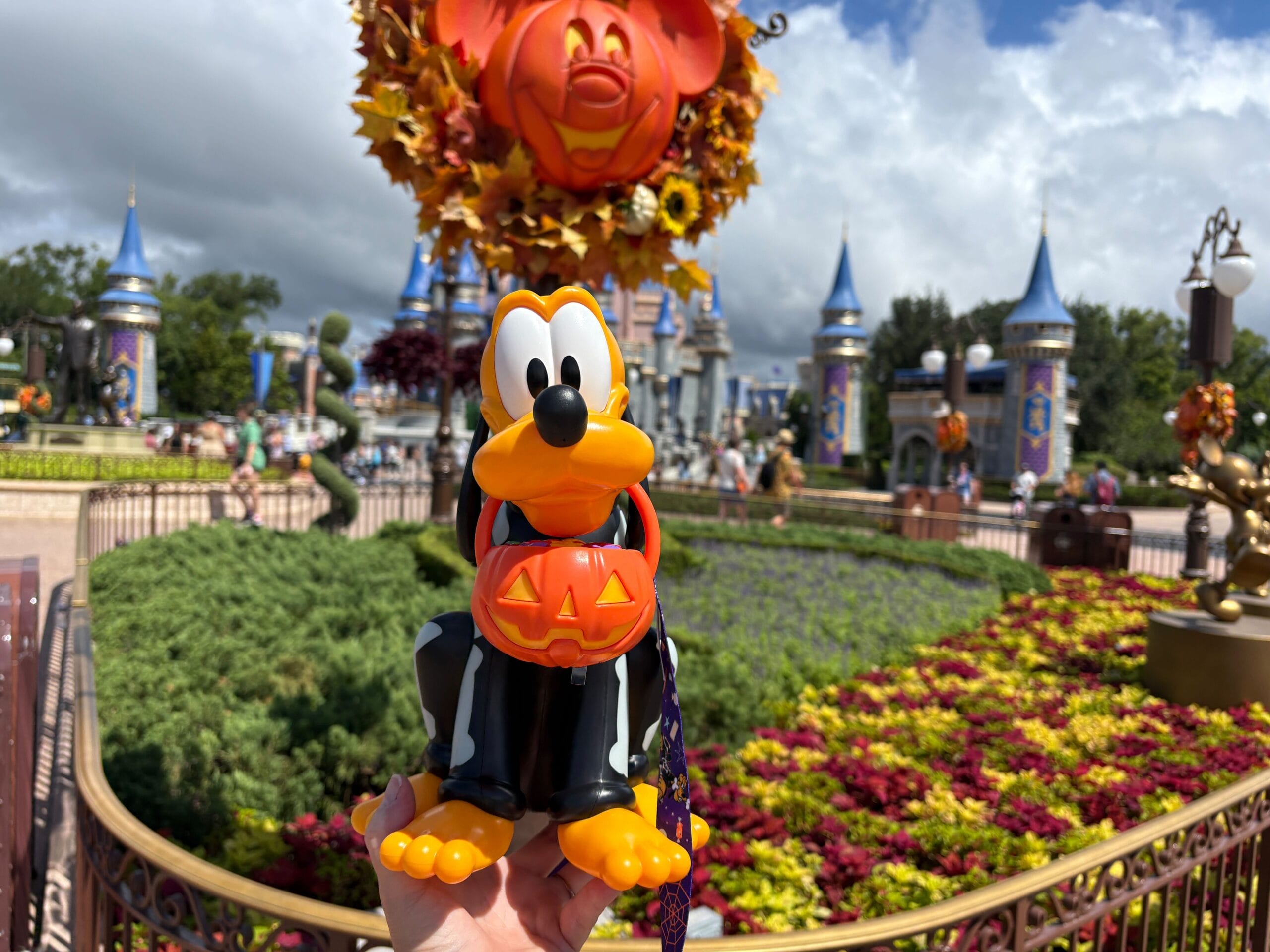A hand holds a Halloween Pluto popcorn bucket at Disney World with festive fall decor and castle turrets in the background.