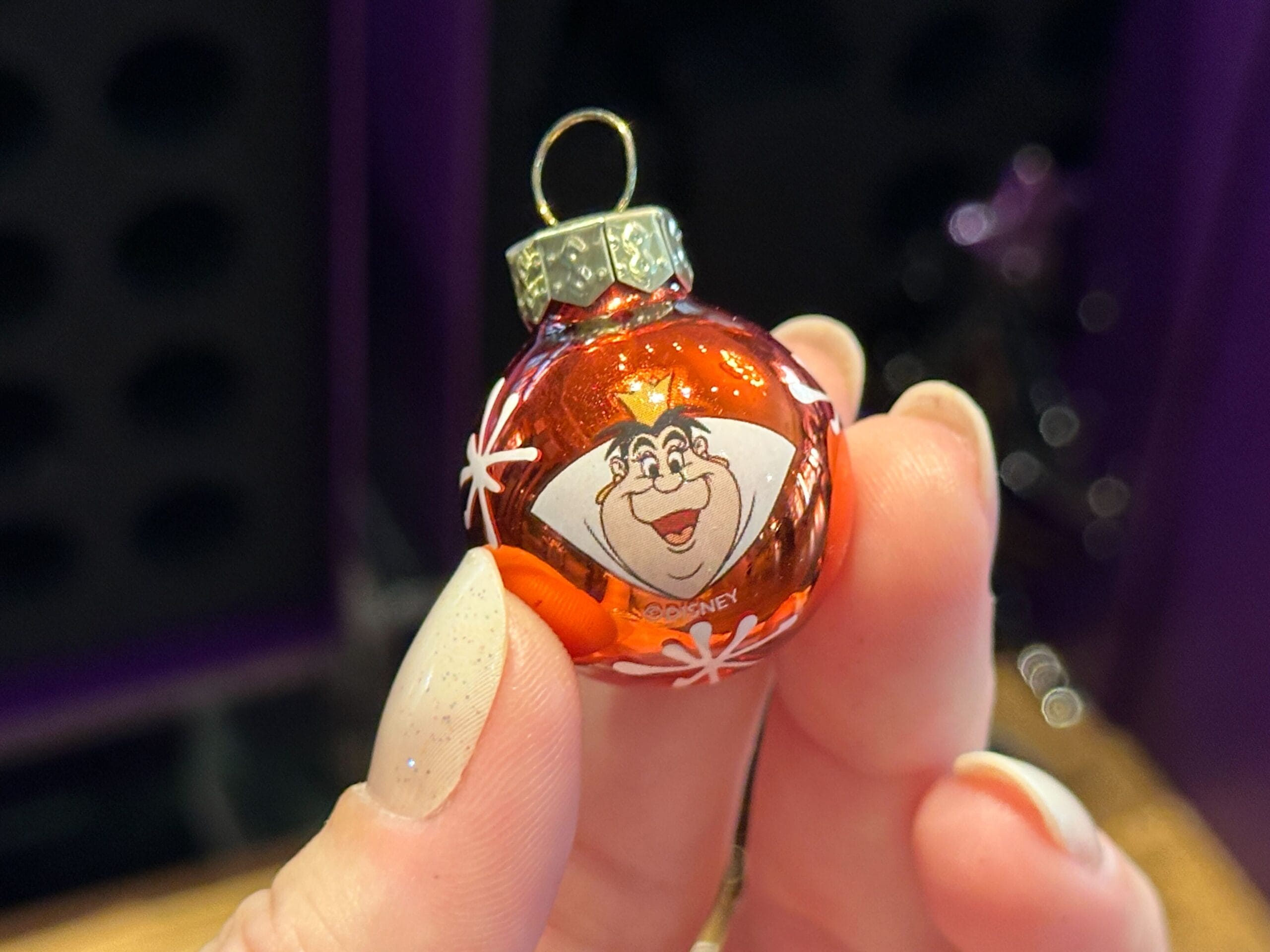 A hand holding a red Villain Ornament with a cartoon face, white decorations, and a gold loop on top.